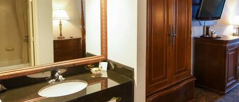 Un baño con un espejo grande, un lavabo, un tocador con cajones, un bote de basura y un televisor montado en la pared.