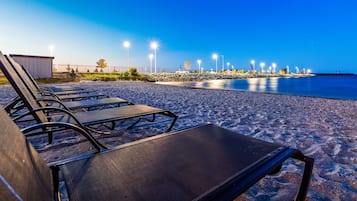 A beach with sun loungers and a poolside area, surrounded by a stone wall and street lamps.