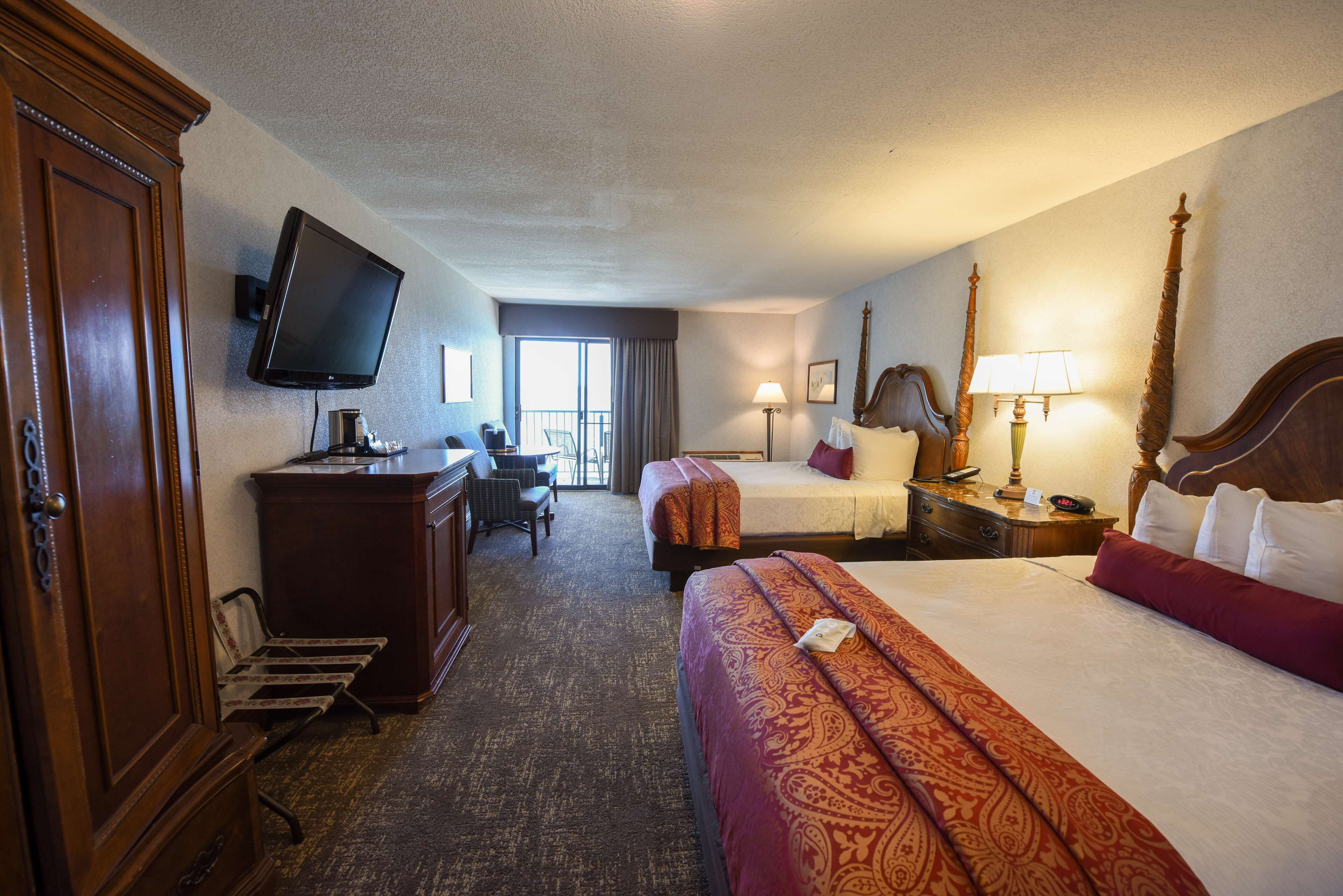 A hotel room with a large bed, a smaller bed, a desk with a chair, a television mounted on the wall, and a window with curtains.