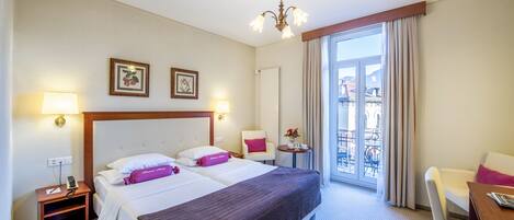 A hotel room with two beds, a desk, a chair, a balcony view, and a chandelier.