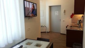 A hotel room with a television mounted on the wall, a small table with a remote control, a kitchen area with a sink and a kettle, and a door leading to another room.
