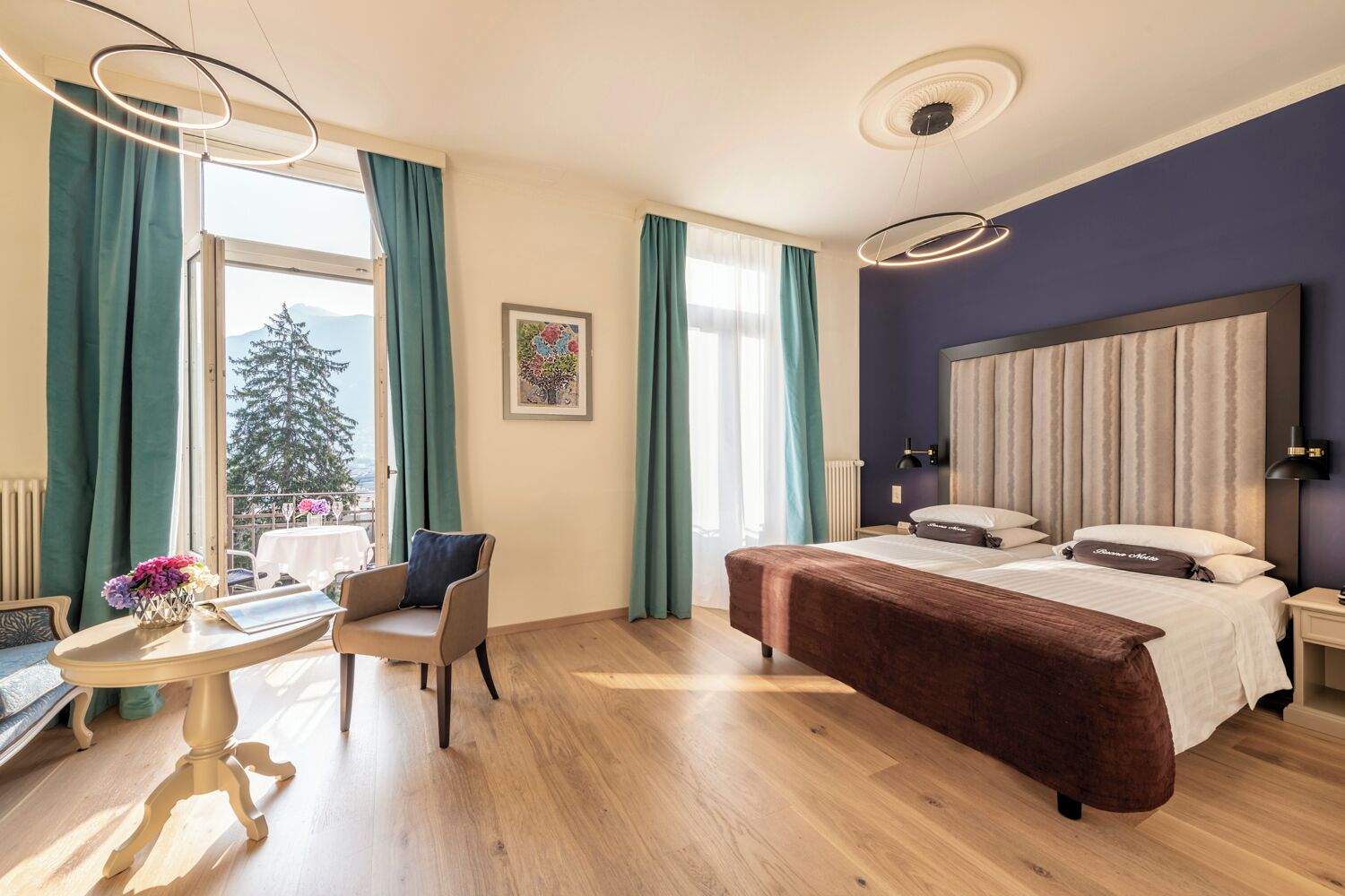 Una habitación de hotel con una cama, una silla, una mesa, un ventanal con cortinas, un radiador y una lámpara fijada a la pared.