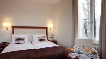 A hotel room with a large bed, bedside lamps, a small table with breakfast items, and a window with a view of trees.