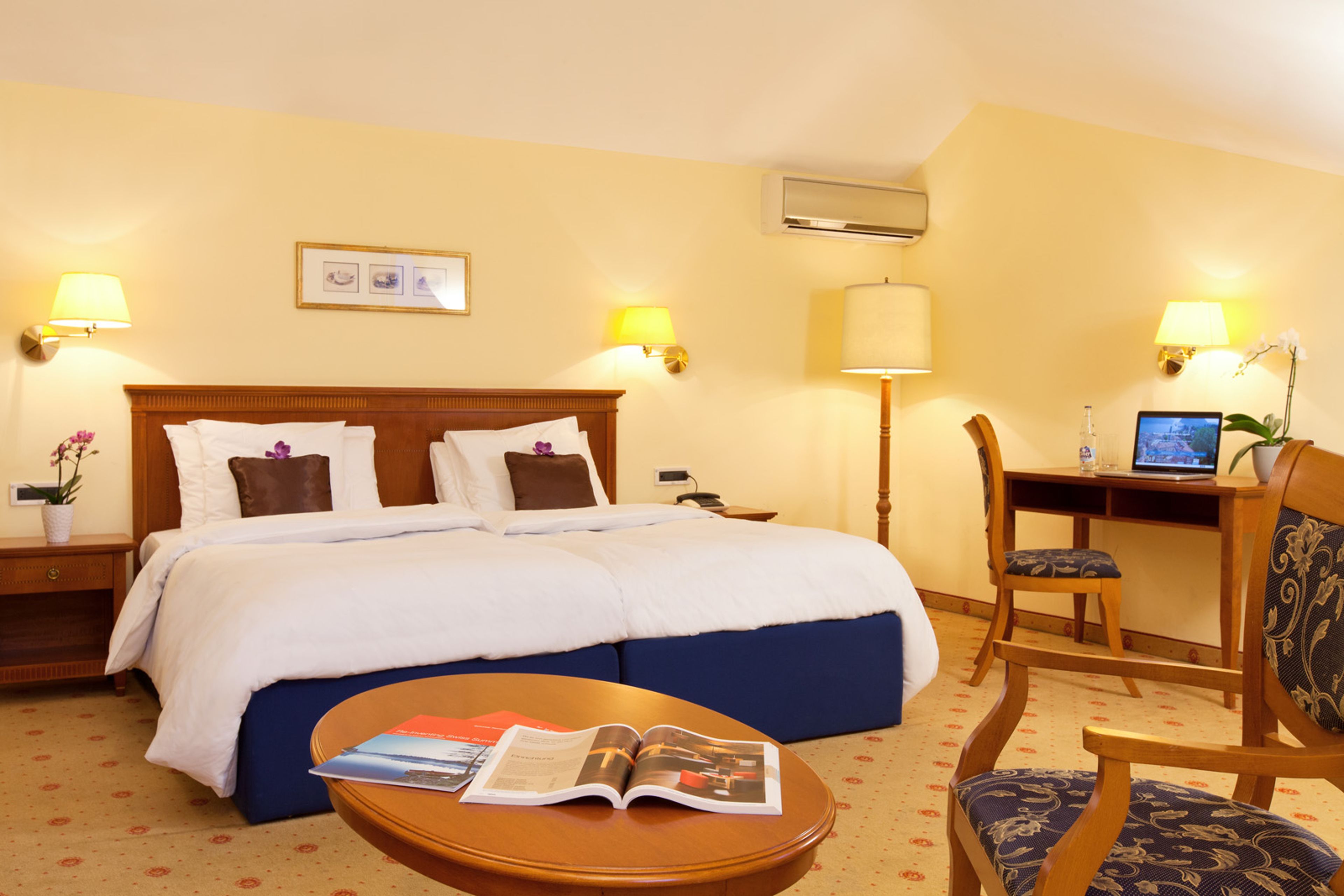 A hotel room with a bed, bedside tables, a desk with a computer, a chair, and a coffee table with a magazine.