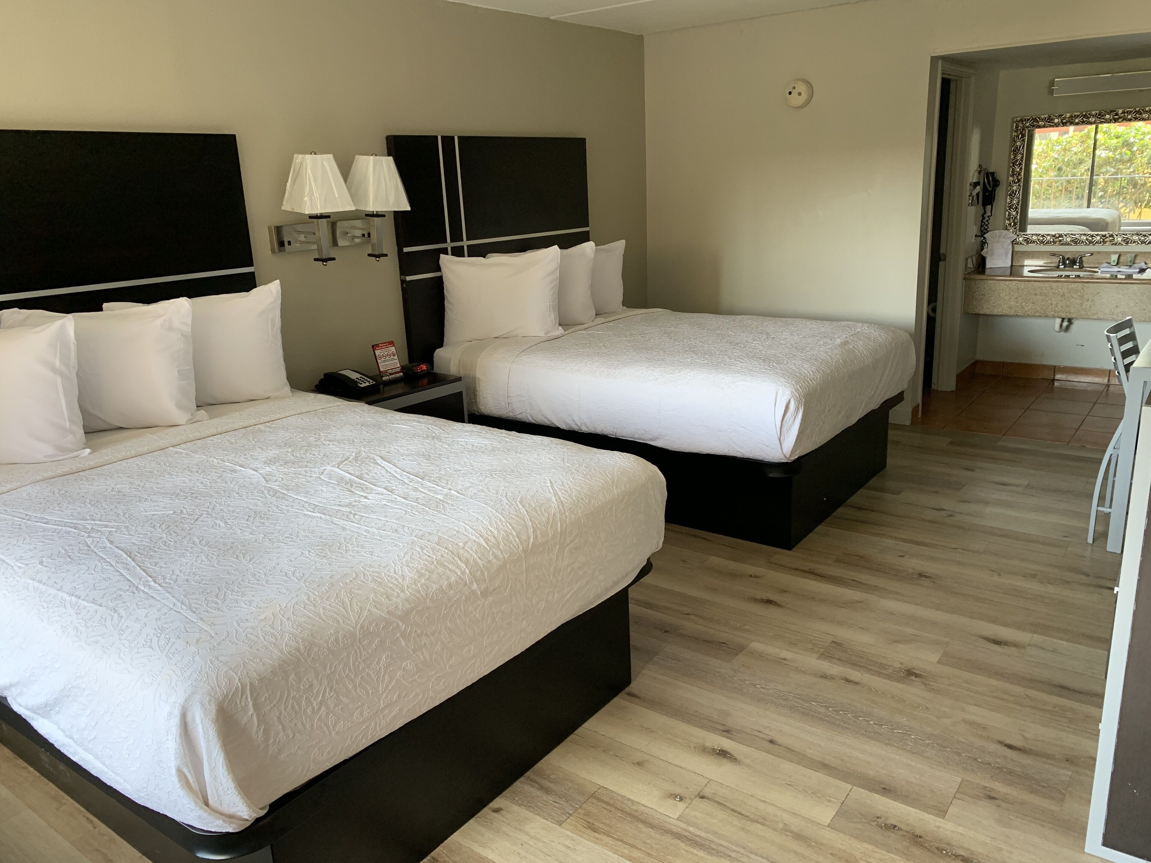 A hotel room with two beds, a nightstand with a phone, wall-mounted lamps, and a bathroom visible through an open door.