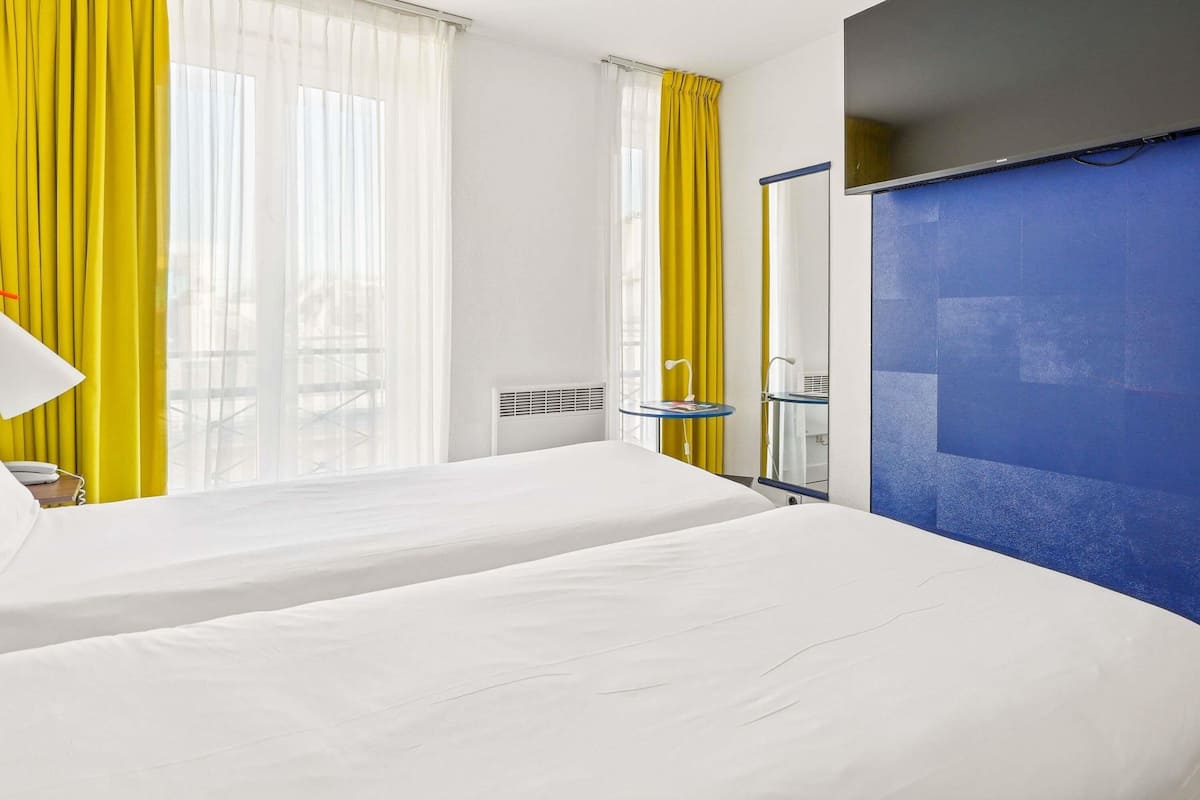 Habitación de hotel con dos camas, un ventanal con cortinas amarillas, una pared de acento azul y una mesita de noche pequeña con una lámpara.