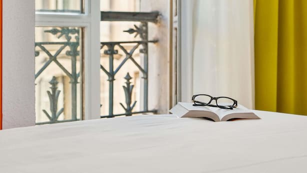 A window with a view of a building, a balcony with metal railings, and a table with a book and glasses. - ibis Styles Paris République (Paris)