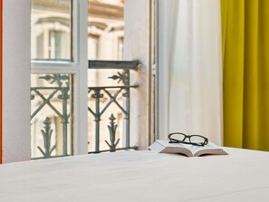 Une fenêtre donnant sur un immeuble, un balcon avec des garde-corps en métal et une table sur laquelle se trouvent un livre et des lunettes.