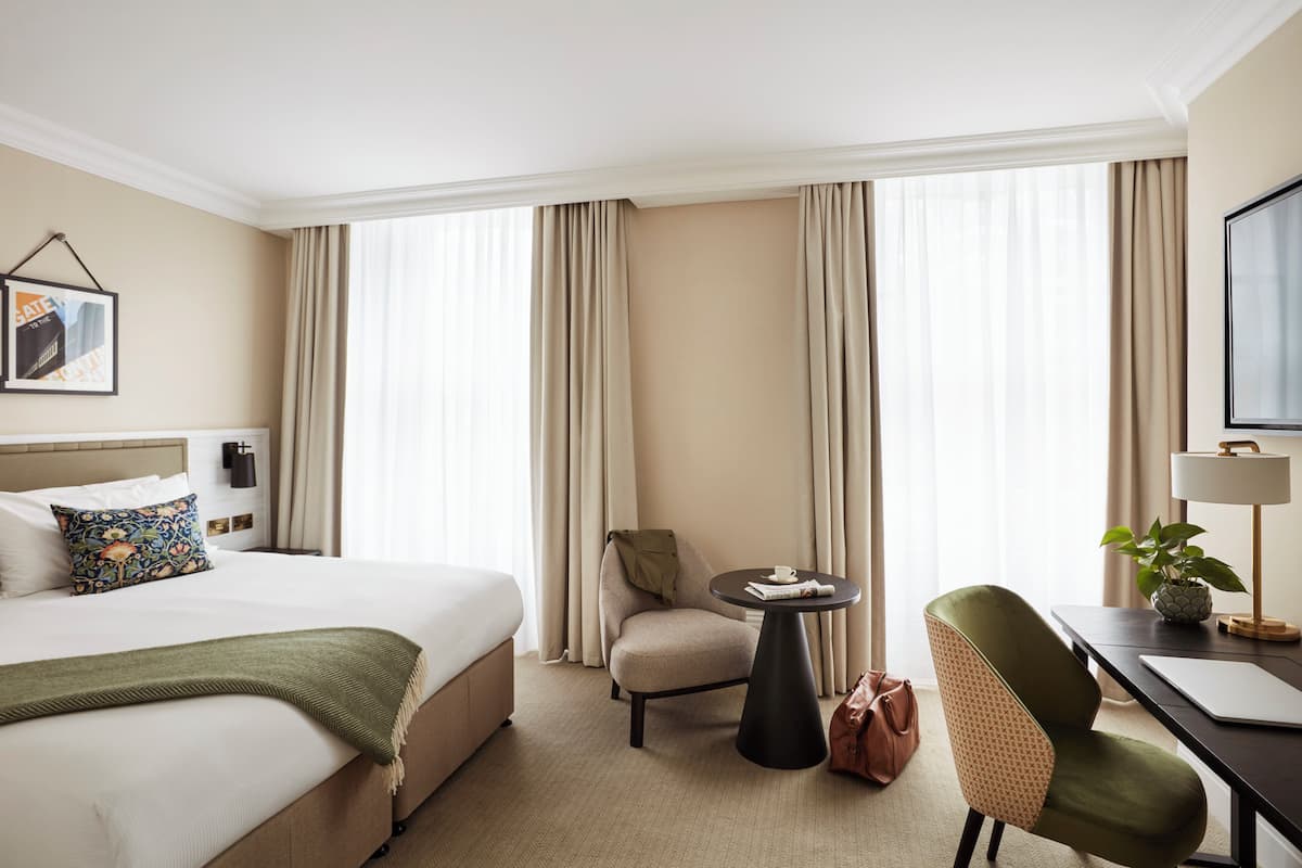 A hotel room with a bed, a chair, a small table, a desk, a lamp, and a plant.