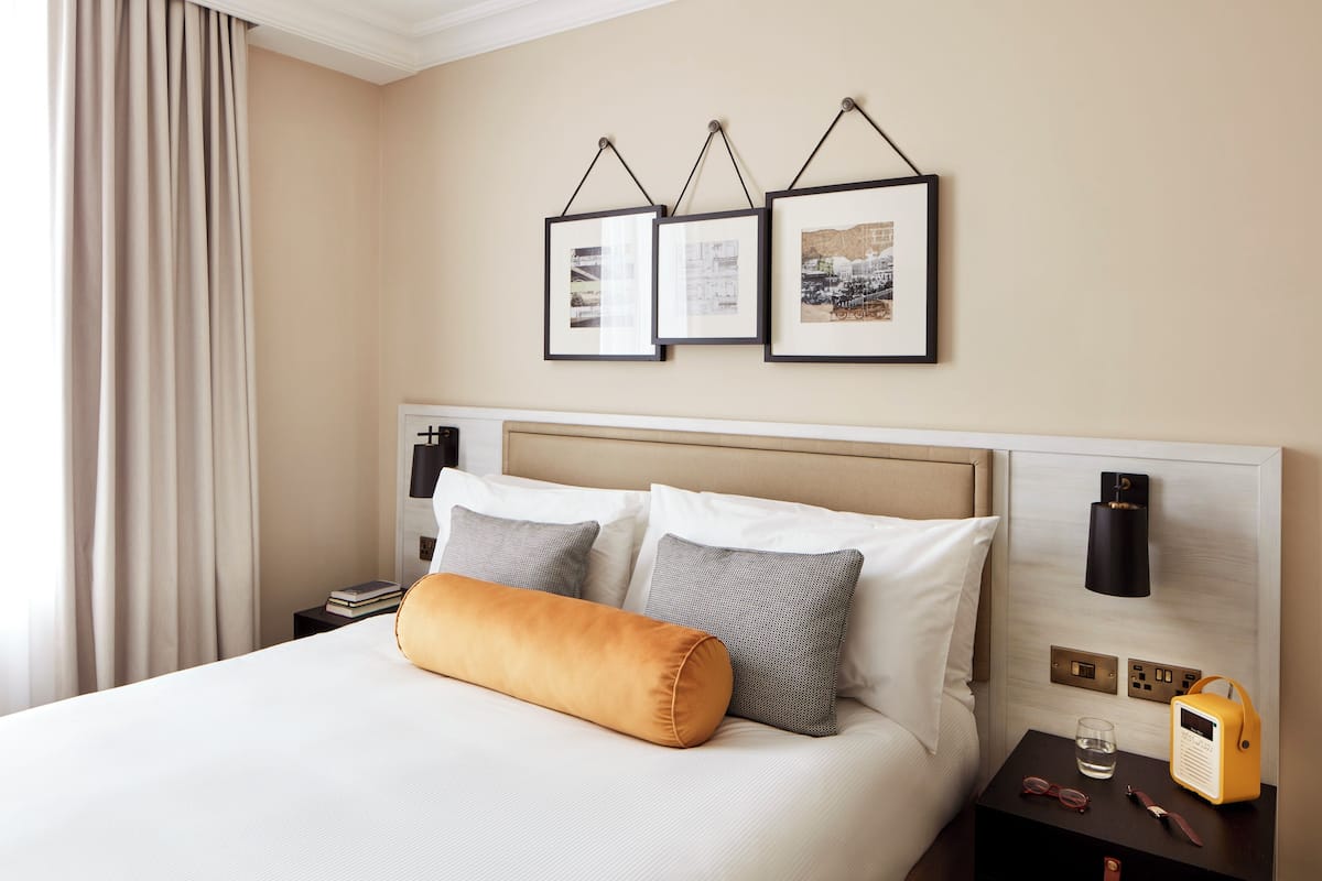 A neatly made bed with pillows, a bedside table with a radio, and framed pictures on the wall.