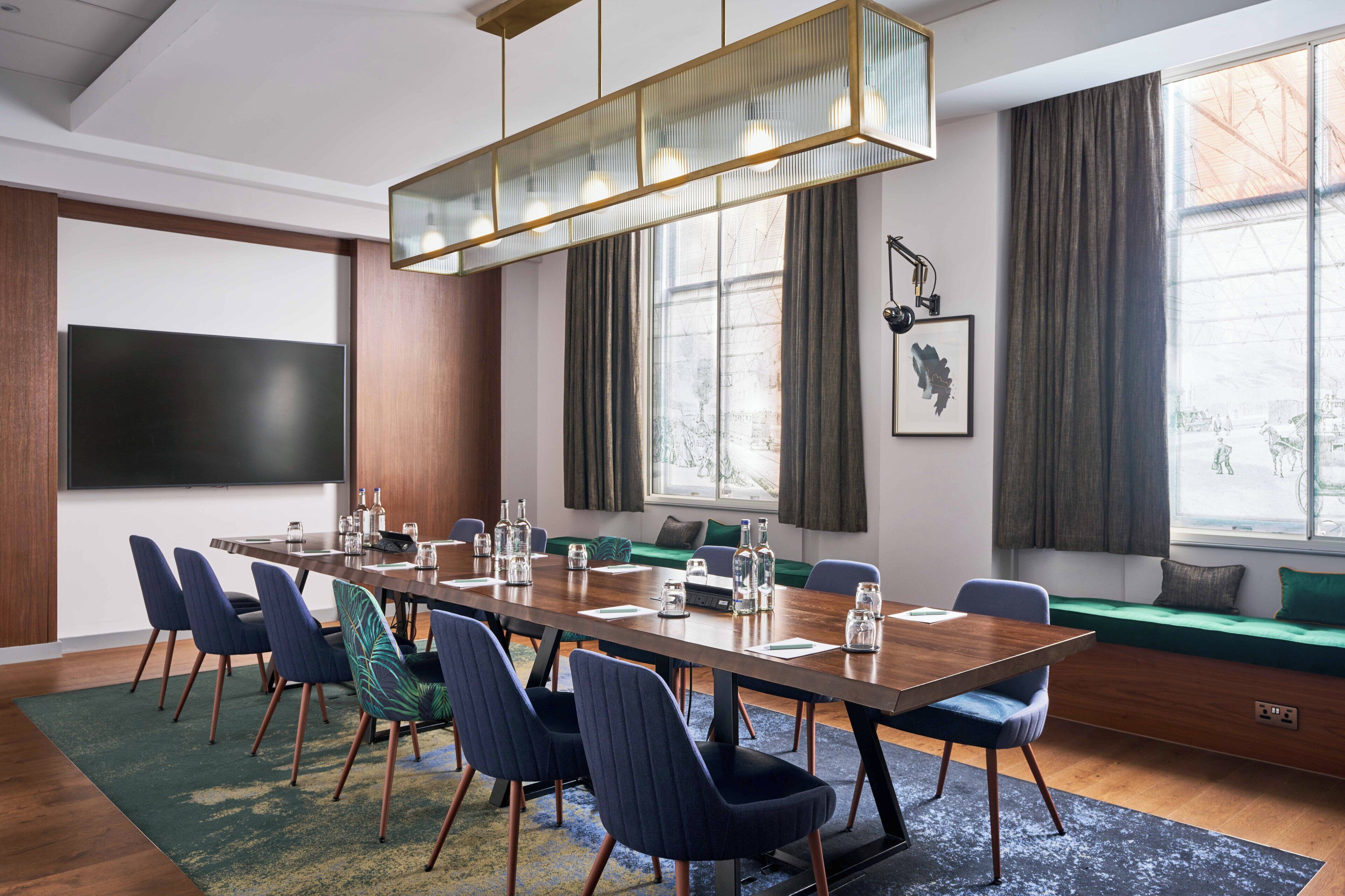 A conference room with a long table, chairs, a large screen, and a window with curtains.