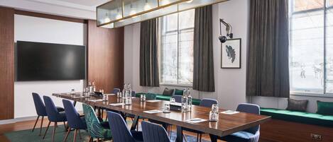 A conference room with a long table, chairs, a large screen, and a window with curtains.