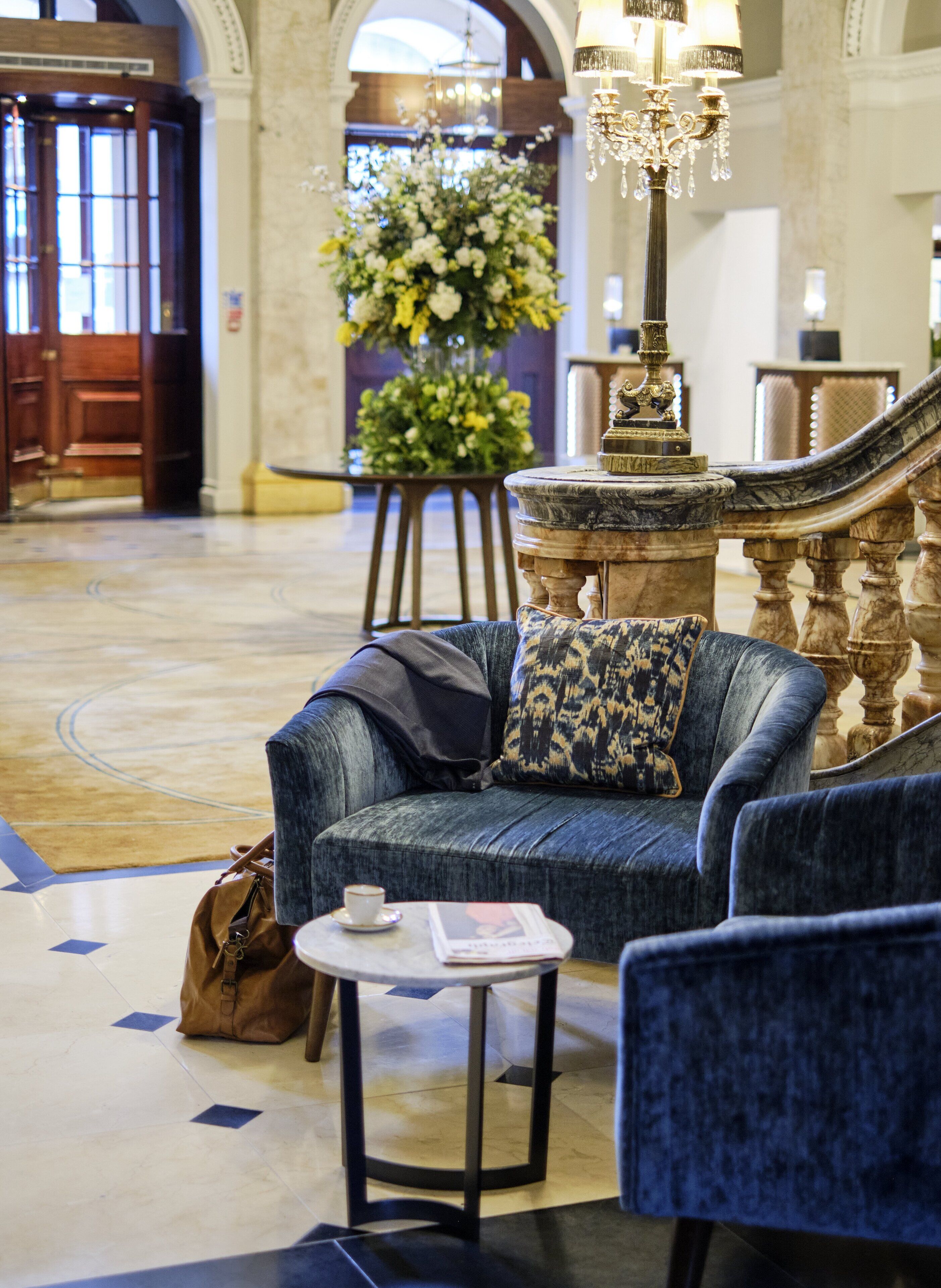 Un hall d’entrée avec un canapé bleu, une petite table sur laquelle se trouvent une tasse et un magazine, un grand arrangement floral et un imposant escalier.