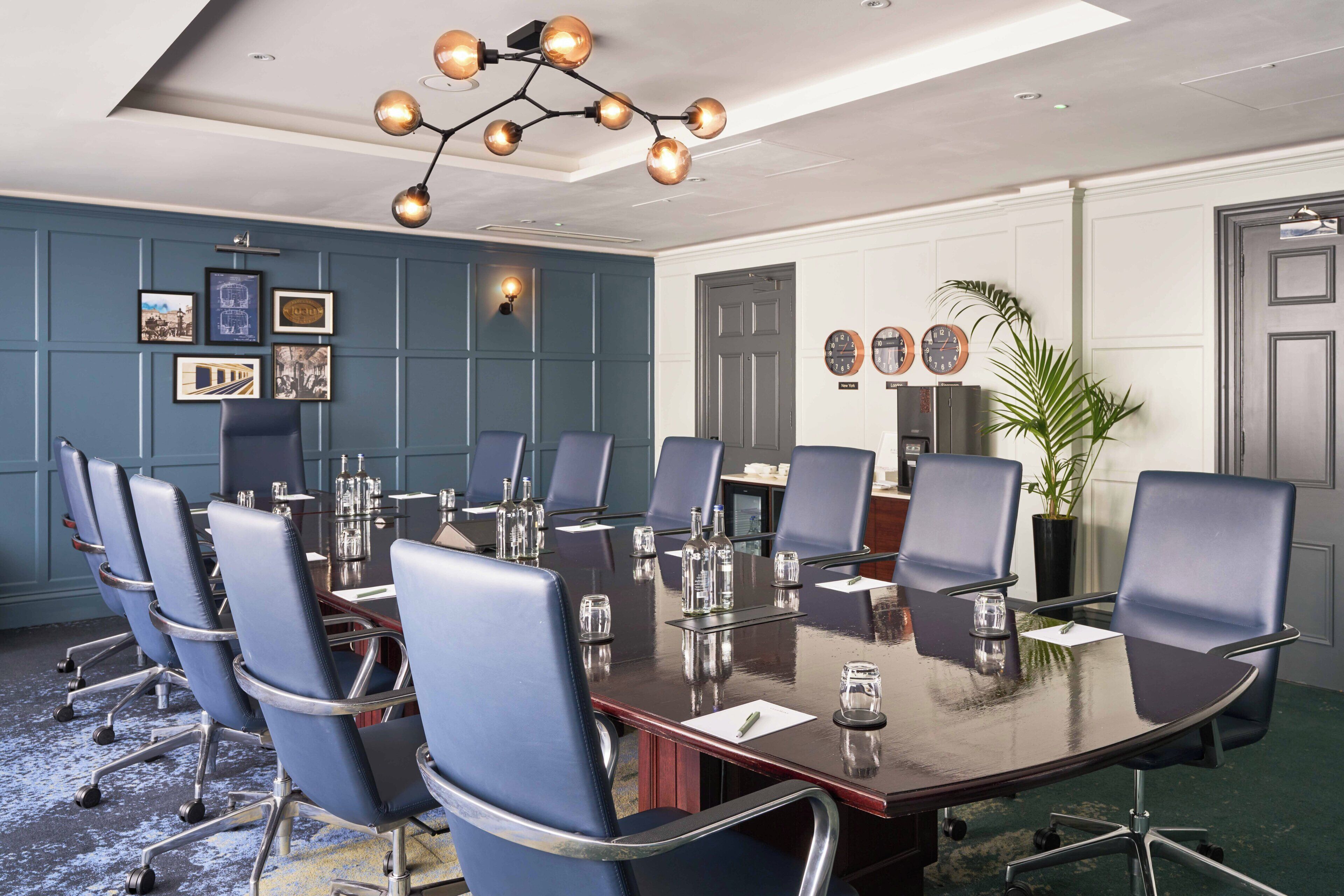 A conference room with a large table, multiple chairs, a chandelier, framed pictures on the wall, and a potted plant.