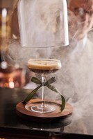 Un cóctel con una copa transparente y un posavasos de madera, rodeado de vapor.