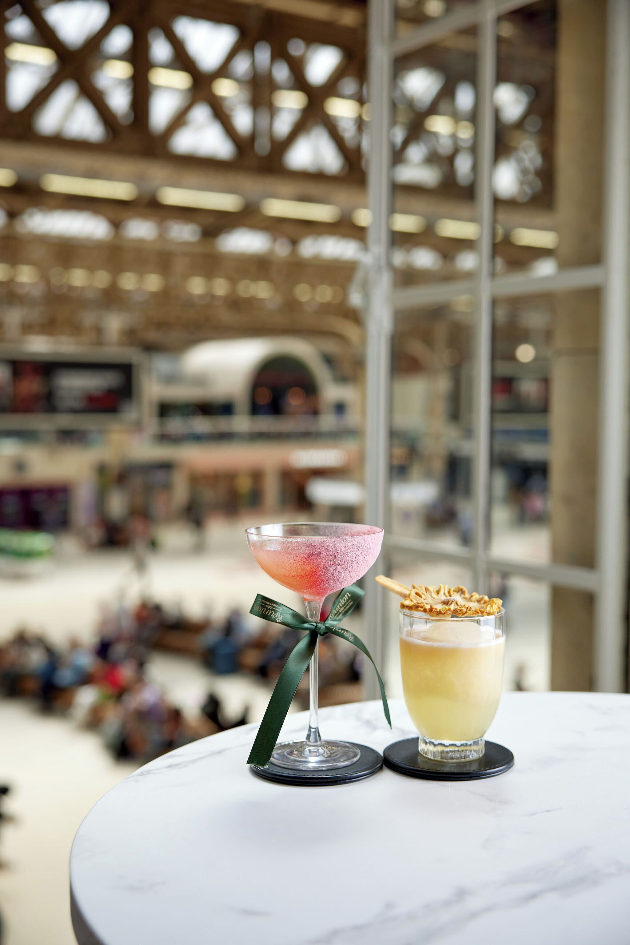 A bar area with two cocktails on a marble table, a large window with a view of a busy shopping area, and a high ceiling with wooden beams.