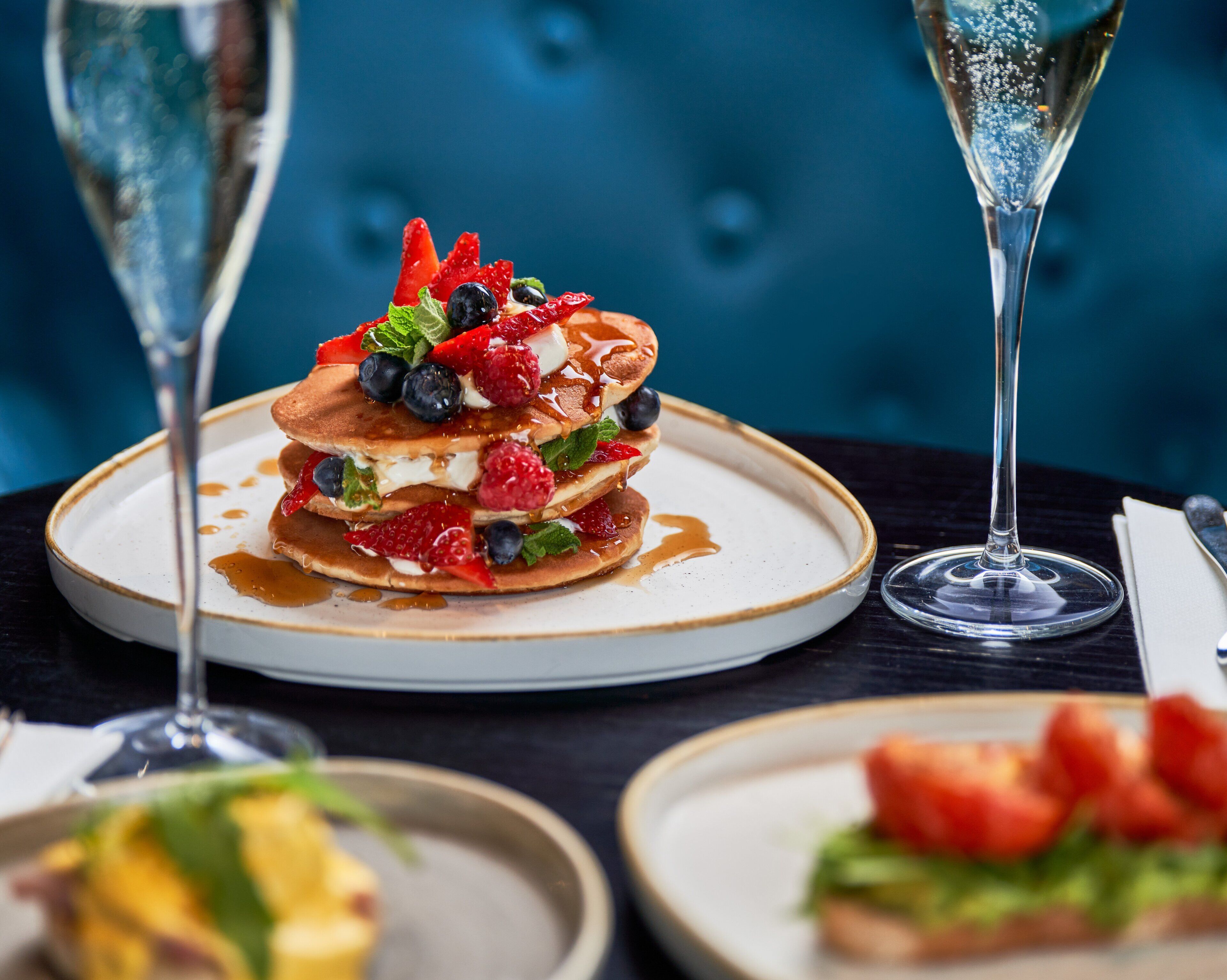 A dessert plate with pancakes topped with berries and syrup, accompanied by two champagne glasses on a table.