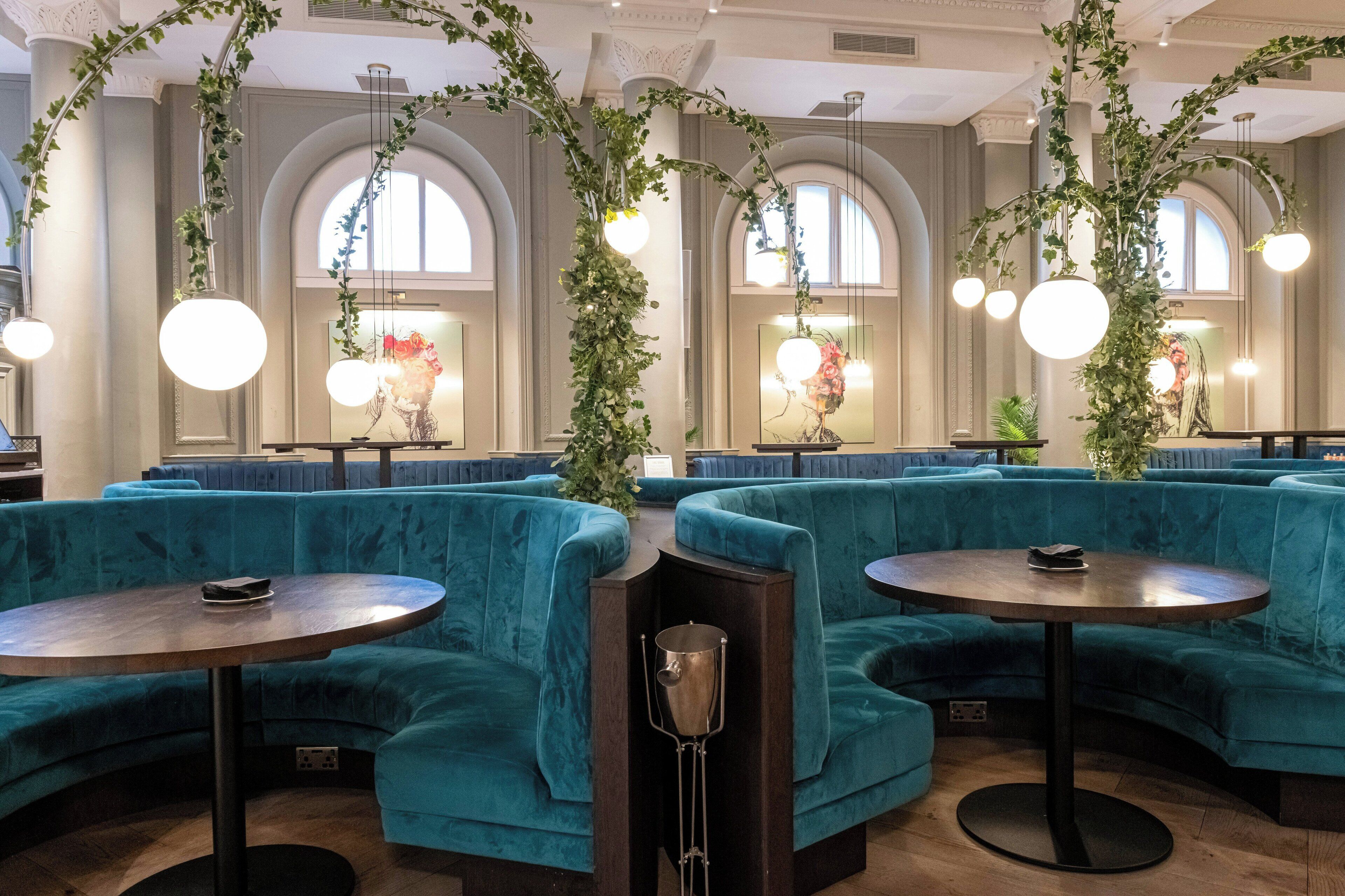 A restaurant with teal upholstered seating, round tables, and hanging plants.