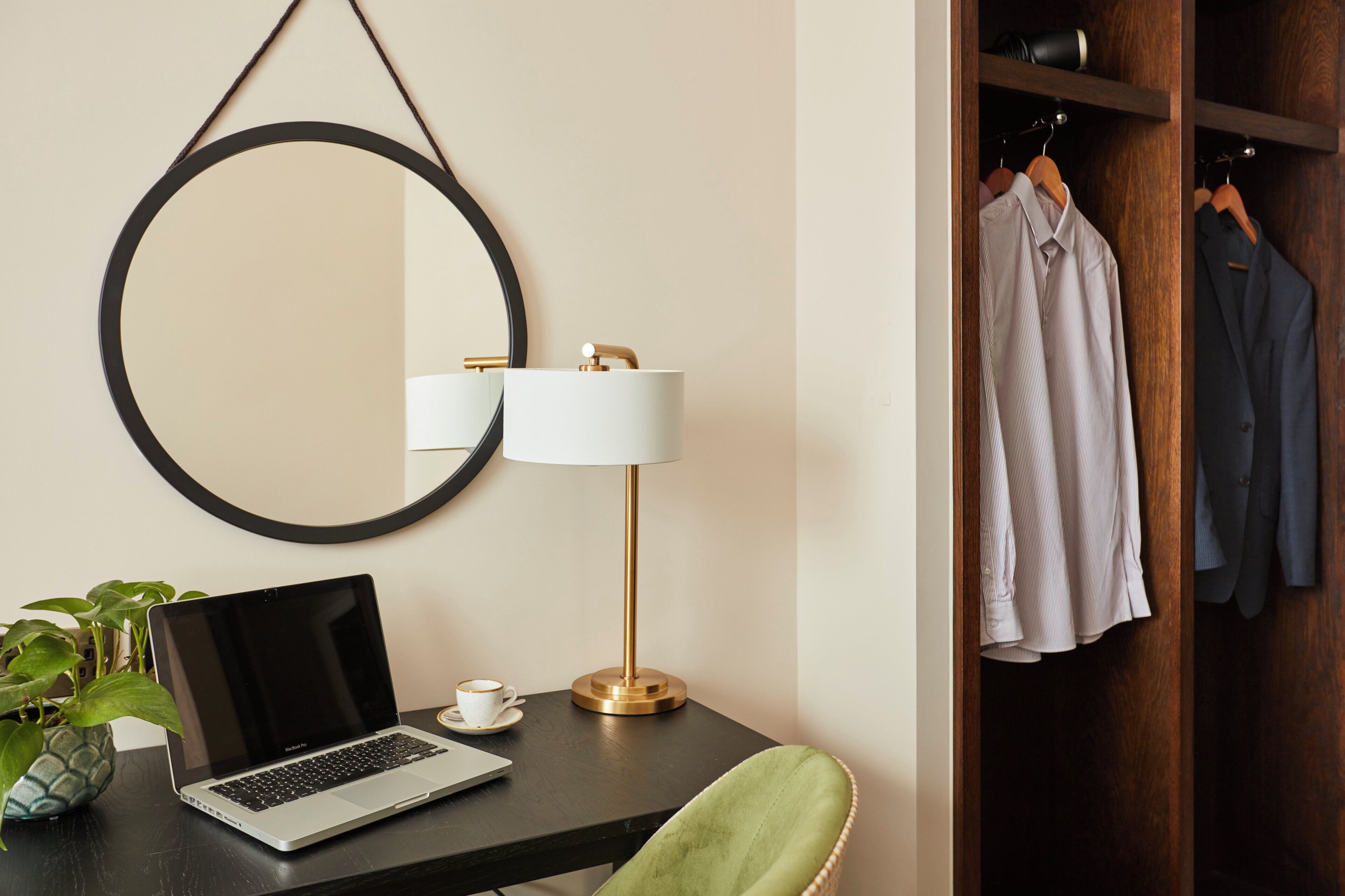 A hotel room with a desk, laptop, coffee cup, lamp, mirror, and a wardrobe with clothes.