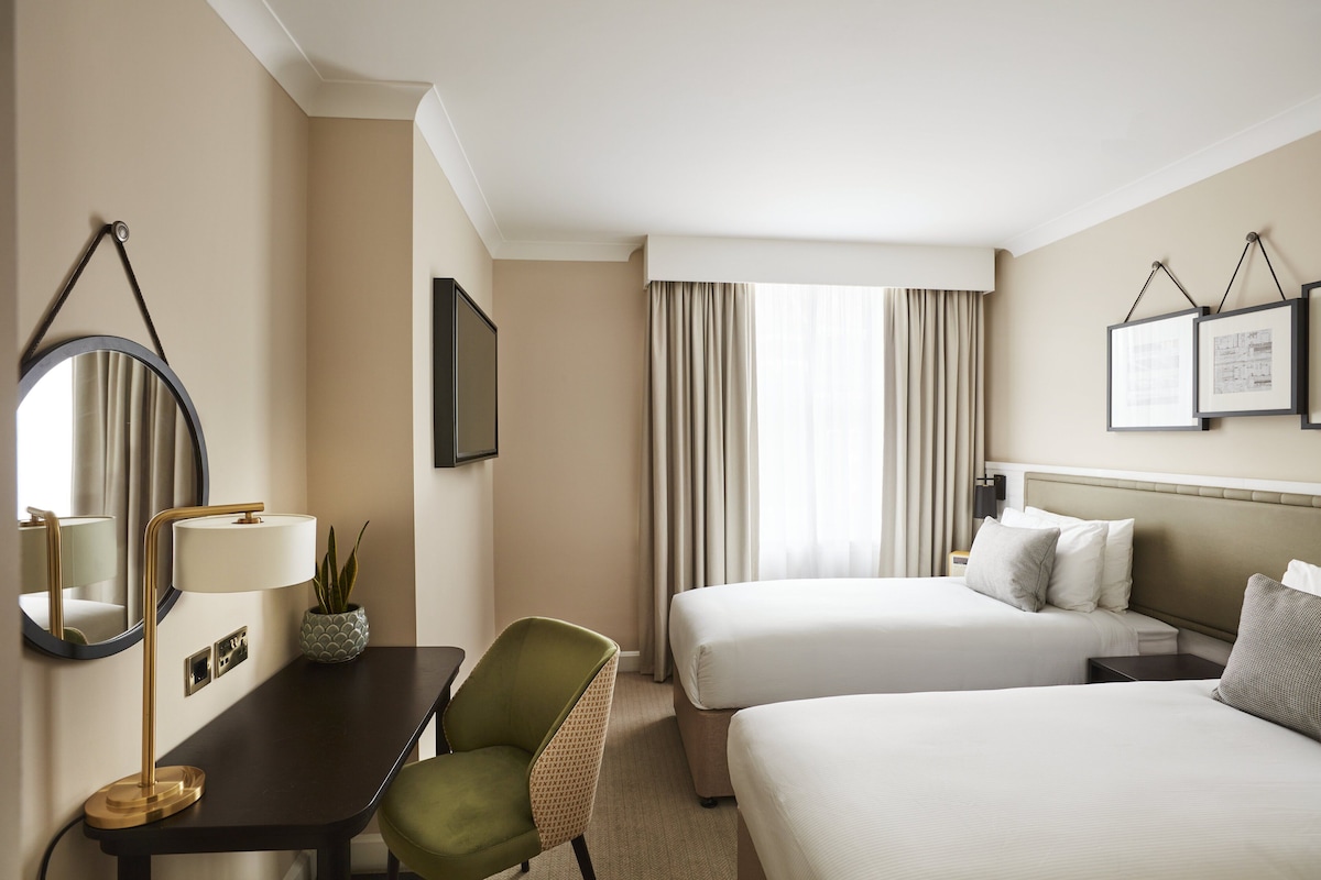 Een hotelkamer met twee bedden, een bureau met lamp, een stoel, een plant in pot en een grote spiegel.