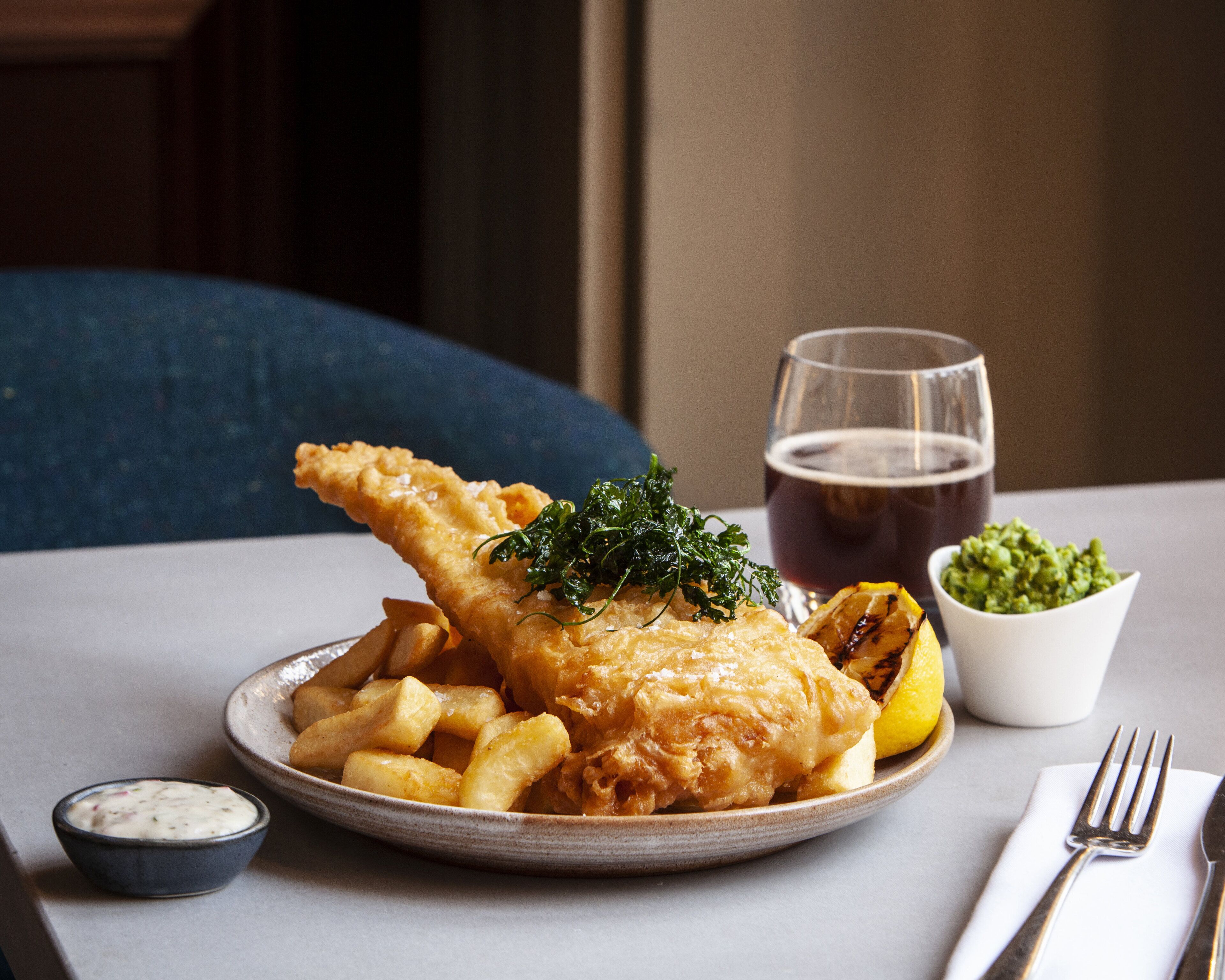 Ein Teller Fish and Chips mit einer Portion Tartarsauce, ein Glas Bier und eine kleine Schale mit Salat auf einem Tisch.