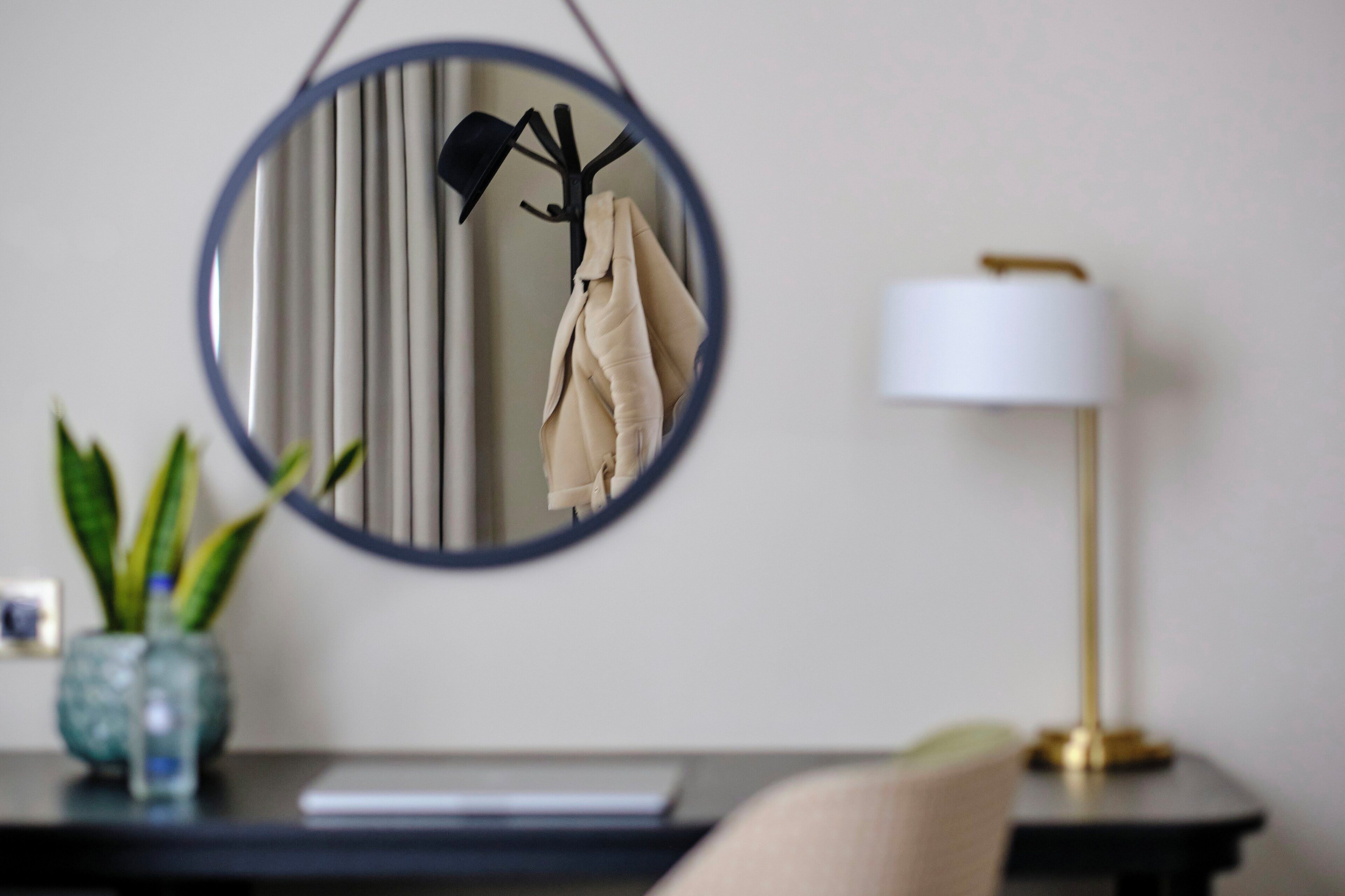 A round mirror with a black frame, a beige coat hanging, a white lamp, and a small plant on a table.