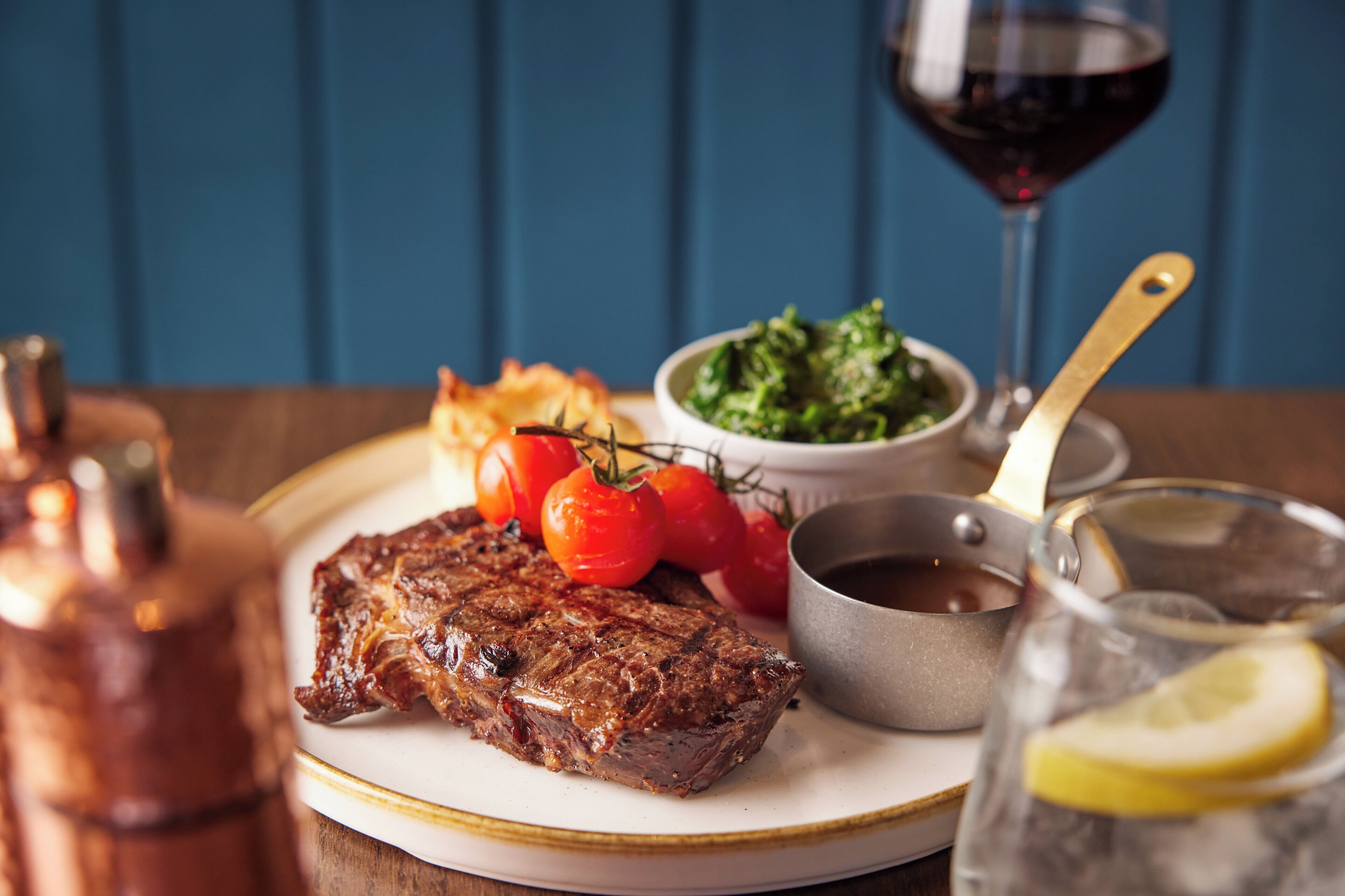 A plate with steak, tomatoes, and greens, a glass of red wine, and a copper pot.