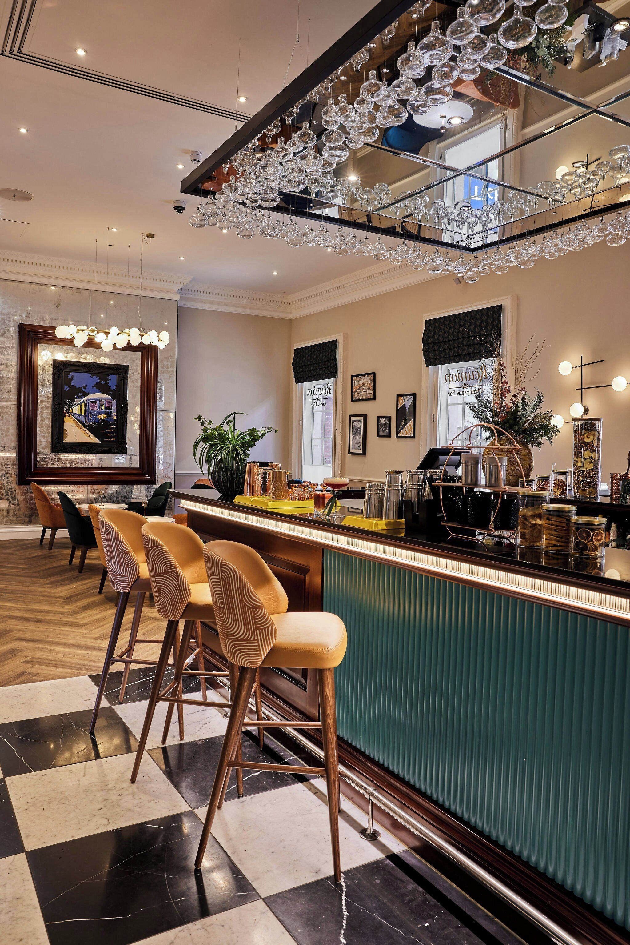 Eine Bar mit Schachbrettmuster-Boden, Barhocker, einer grünen Bar und einer Kronleuchter.