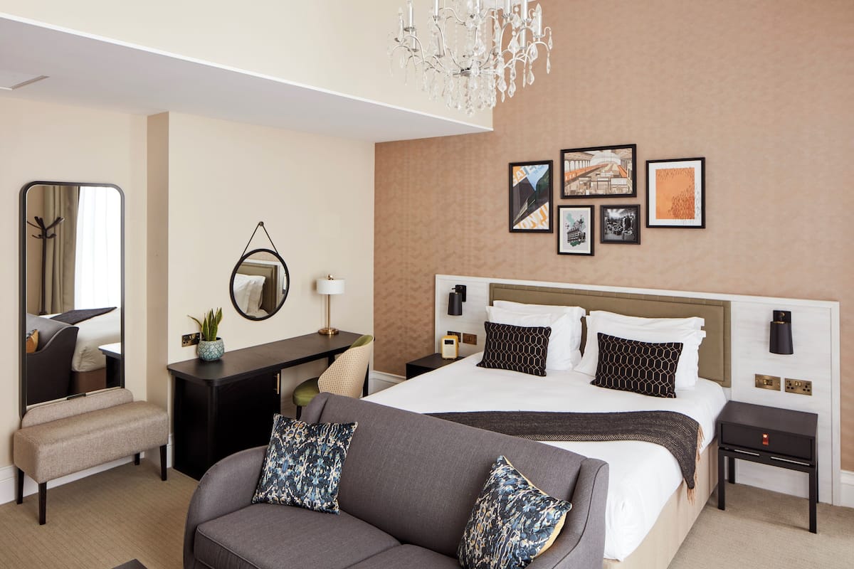 A hotel room with a bed, a desk, a sofa, a mirror, and artwork on the wall.