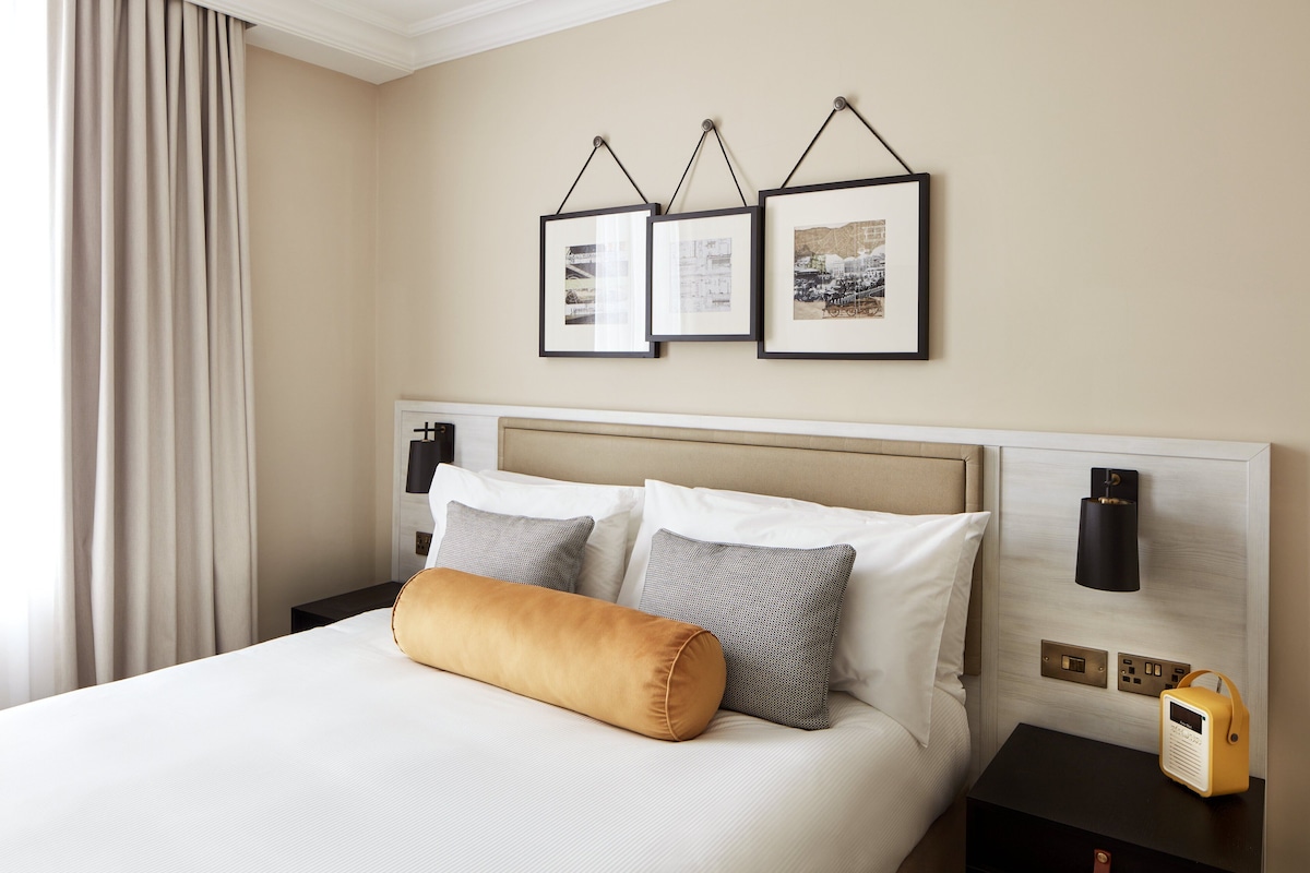 Een keurig opgemaakt bed met hoofdeinde, een nachtkastje met lamp en ingelijste foto's aan de muur.