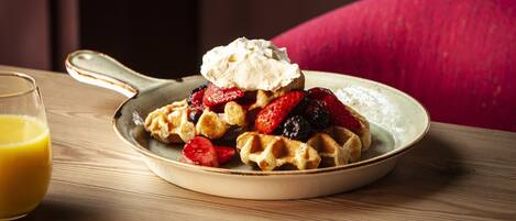 Un plato de waffles cubiertos de crema batida y frutos rojos, junto a un vaso de jugo de naranja sobre una mesa de madera.