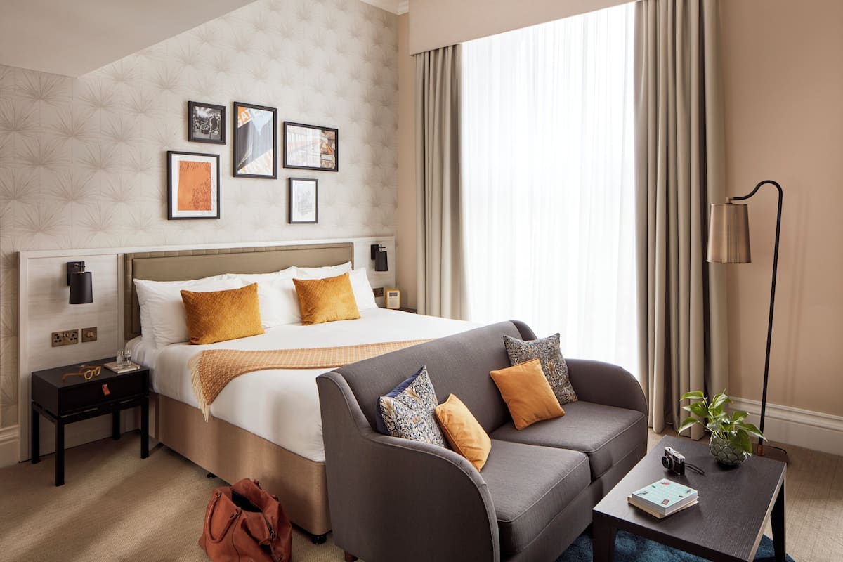 A hotel room with a bed, a sofa, a nightstand, a lamp, and a plant.