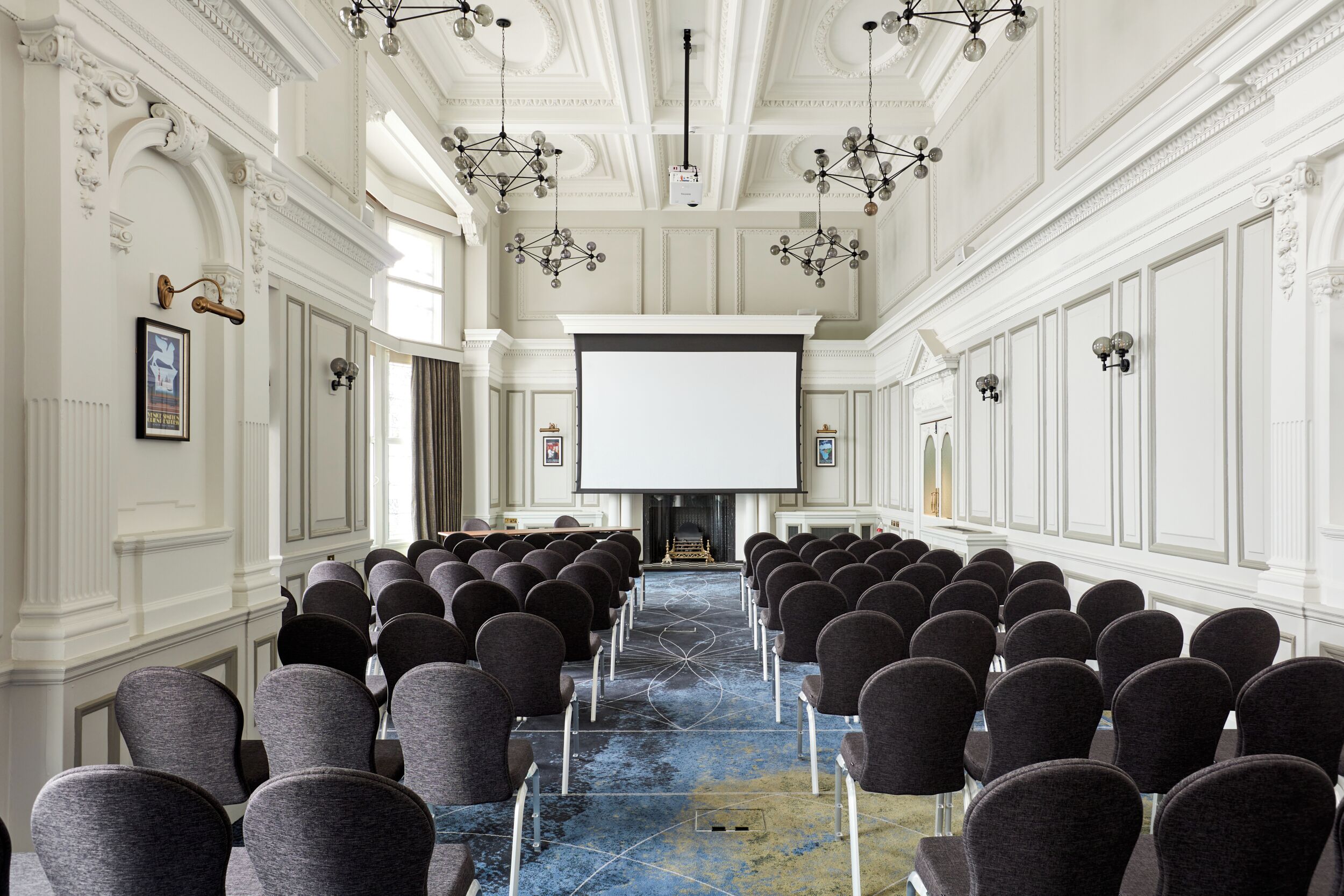 Un salón de conferencias con filas de sillas orientadas hacia una gran pantalla, paredes blancas ornamentadas y candelabros.