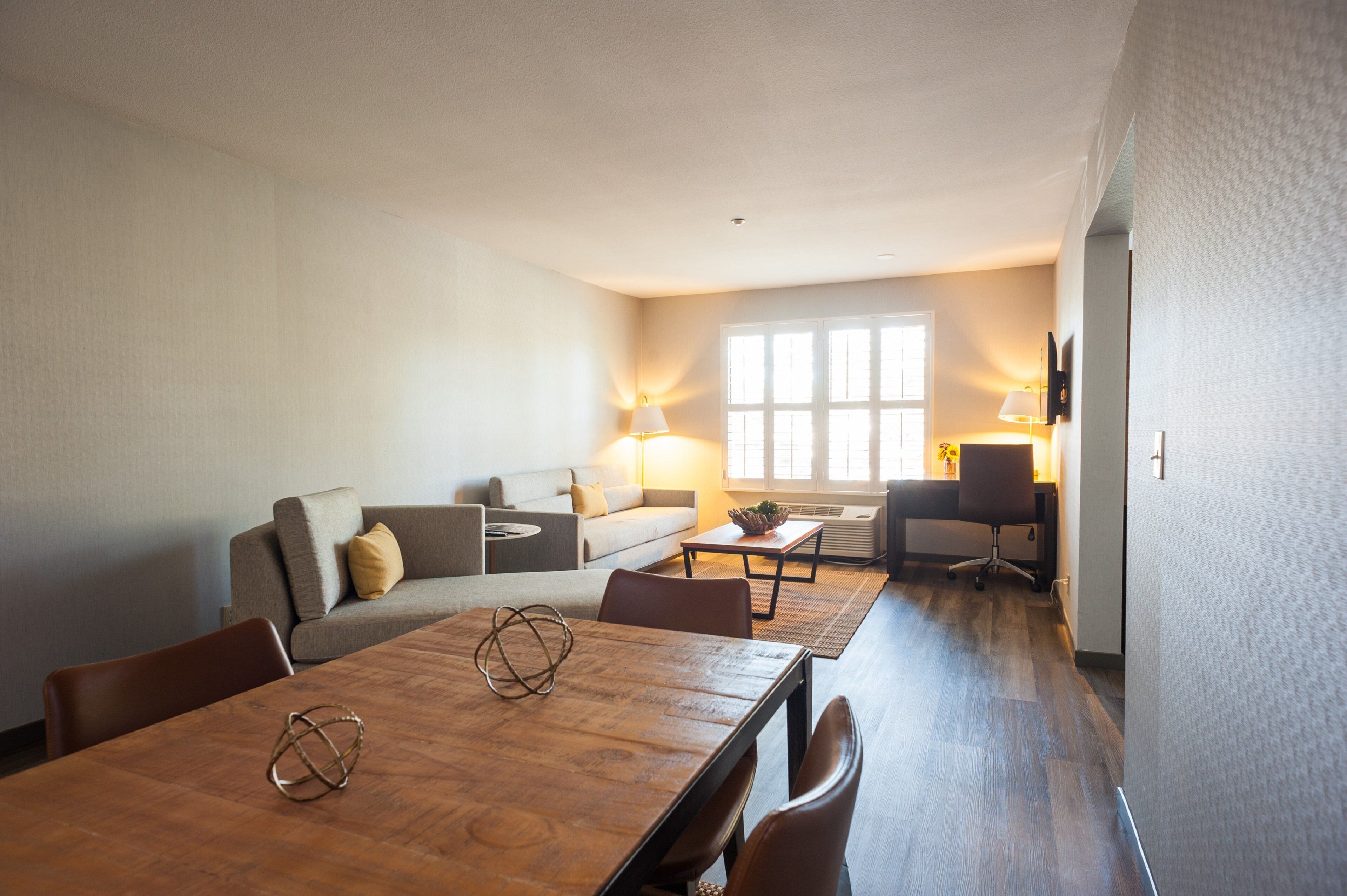 A modern living room with a wooden dining table, a sofa, and a desk with a lamp.
