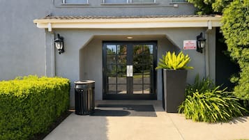 Entrada de edificio con puertas dobles, un basurero, una jardinera con plantas y un letrero de 'No Fumar'.