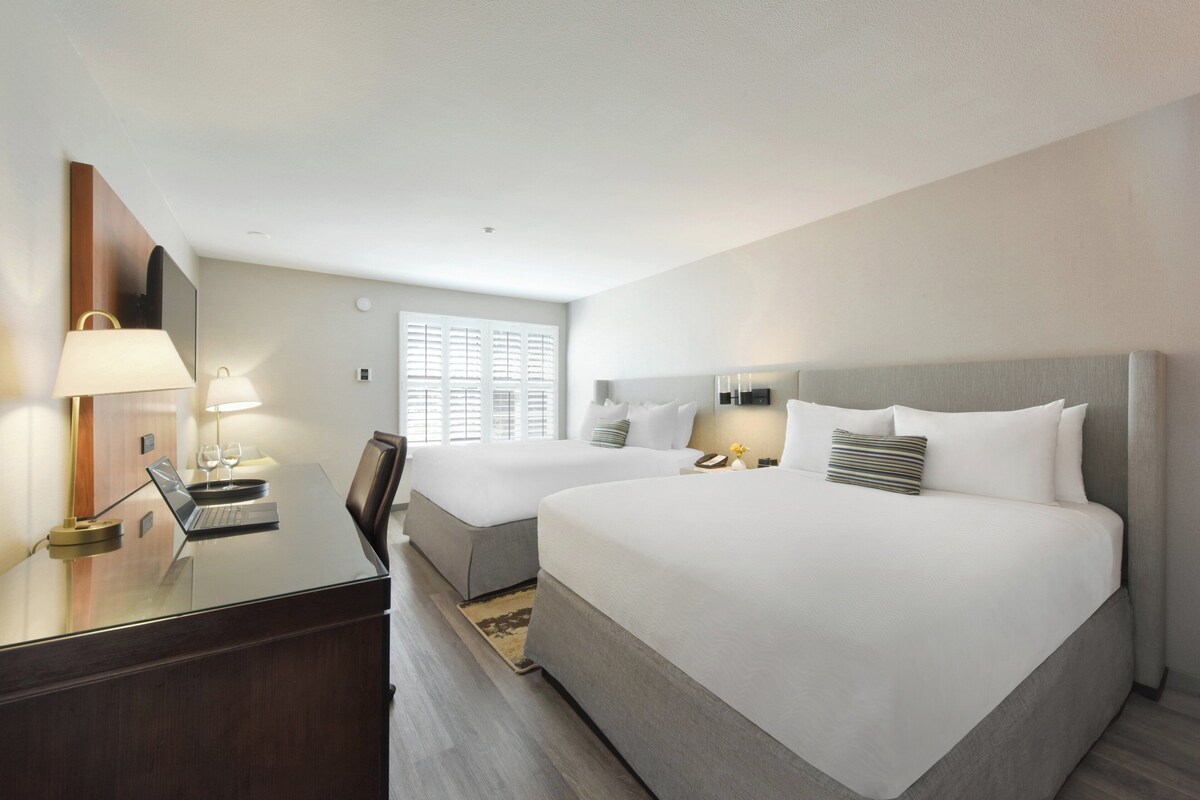 Une chambre d’hôtel avec deux lits, un bureau avec un ordinateur portable et une fenêtre avec des stores.