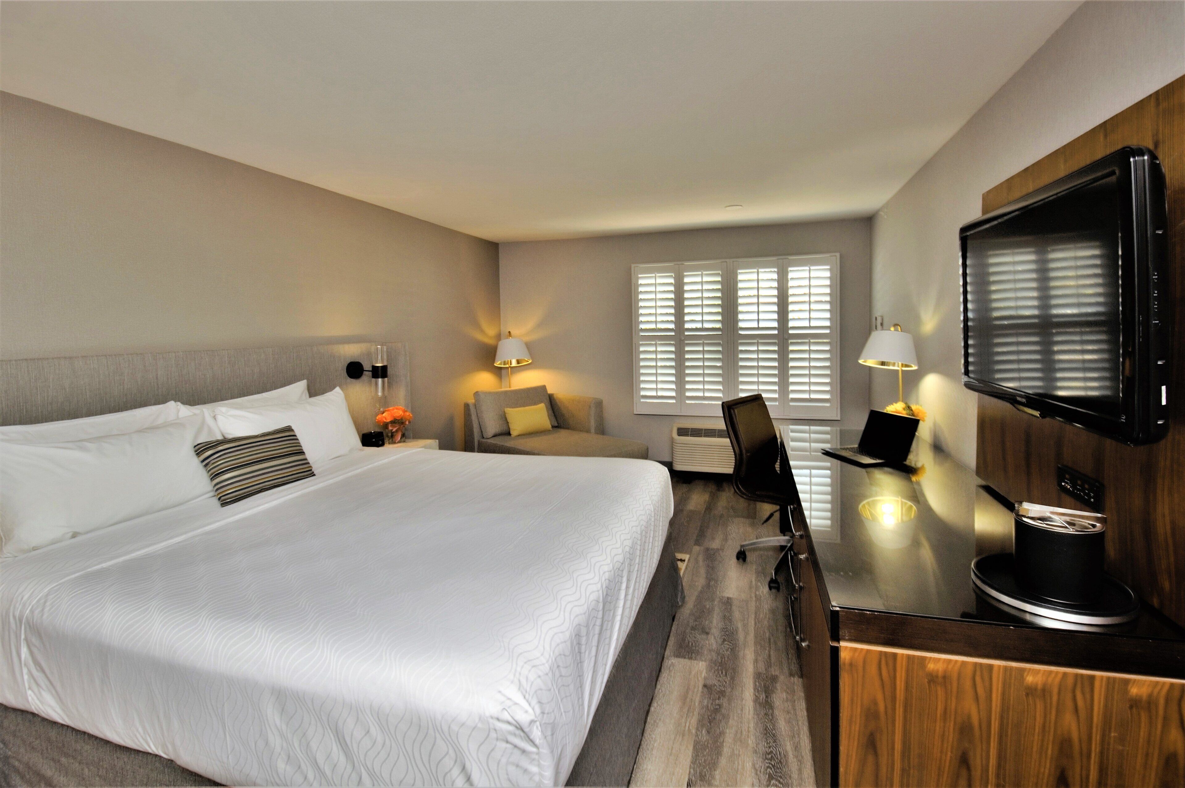 Habitación de hotel con una cama grande, un escritorio con silla, televisión y ventana con persianas.