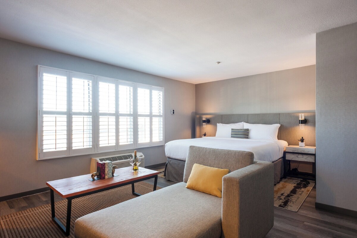 Une chambre d’hôtel avec un grand lit, un canapé, une petite table avec une bouteille, et une fenêtre avec des stores.
