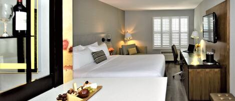 A hotel room with a bed, bedside table, lamp, desk, chair, and a view of the outside through the window.