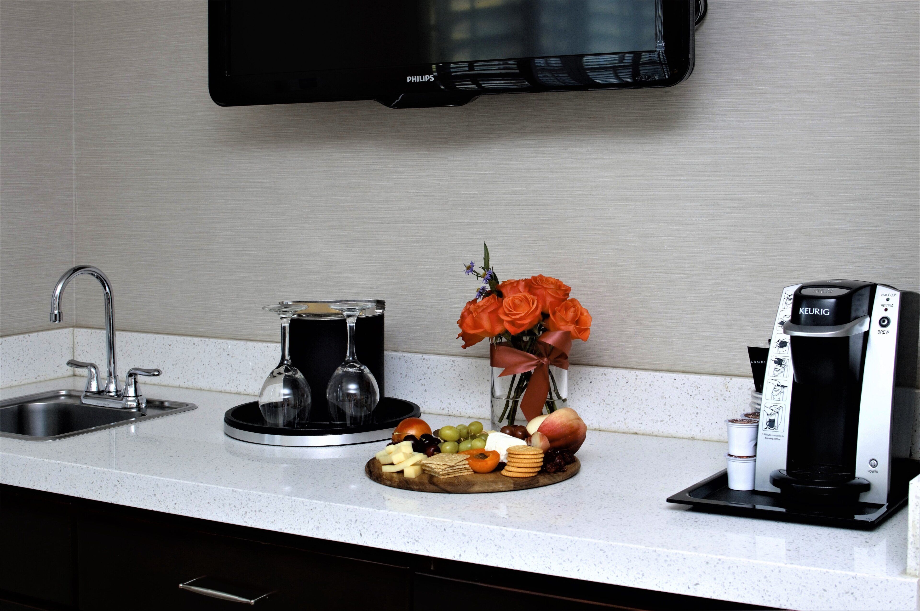 A kitchen counter with a coffee maker, a glass of wine, a vase with flowers, and a cheese platter.