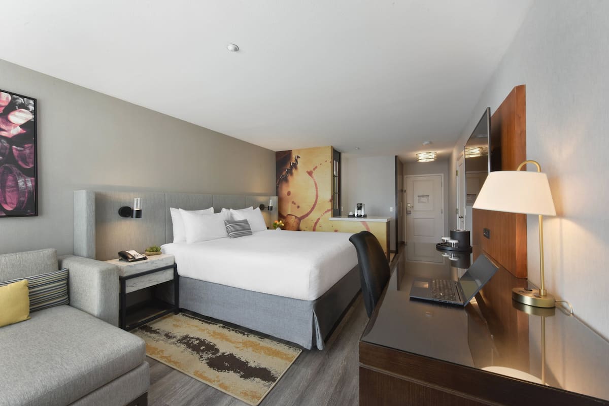 A modern hotel room with a large bed, a desk, a laptop, and a bedside table with a lamp.