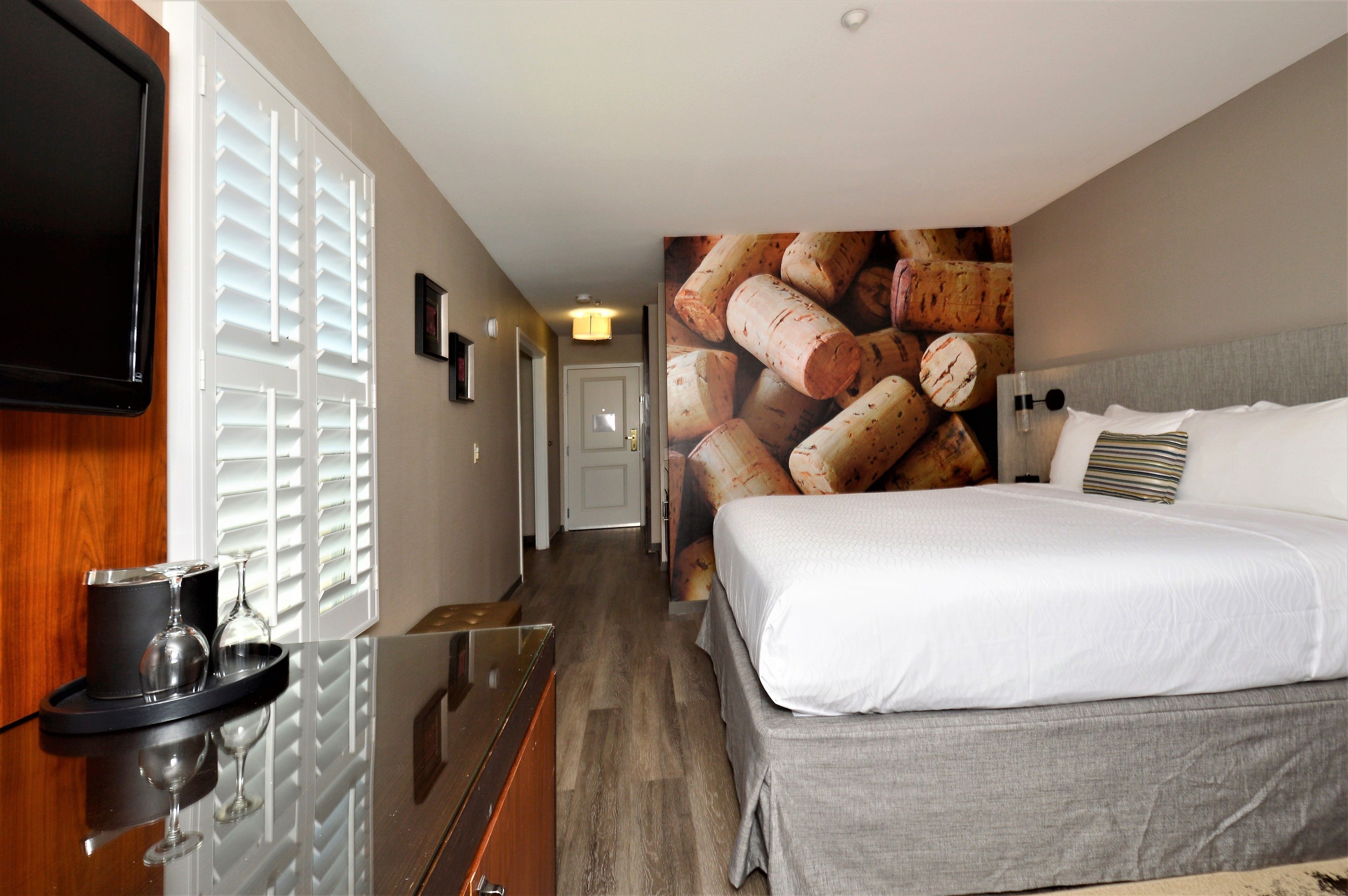 A hotel room with a bed, a TV mounted on the wall, a wine glass and tumbler on a tray, and a wall with a corkscrew image.