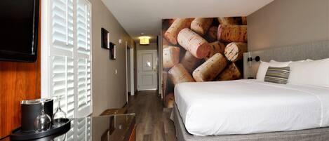 A hotel room with a bed, a TV mounted on the wall, a wine glass and tumbler on a tray, and a wall with a corkscrew image.