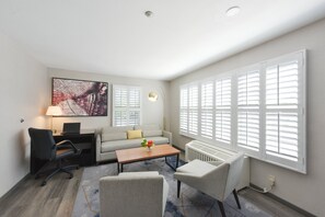 Ein modernes Wohnzimmer mit einer Couch, einem Couchtisch und Sesseln, einem Schreibtisch mit einem Laptop, einer Lampe und einem großen Fenster mit Jalousien.