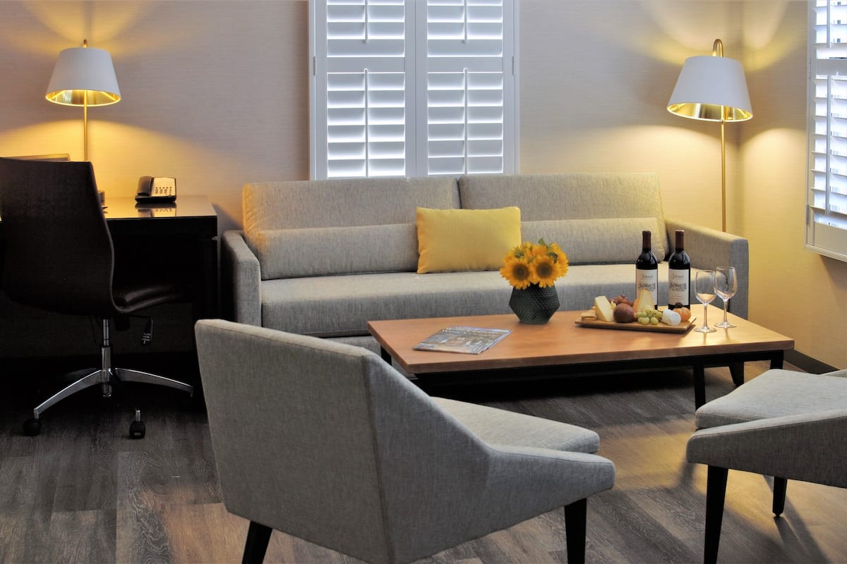 A modern hotel room with a grey sofa, a wooden coffee table with a vase of sunflowers, a dining table with wine bottles and glasses, and a desk with a lamp and a phone.