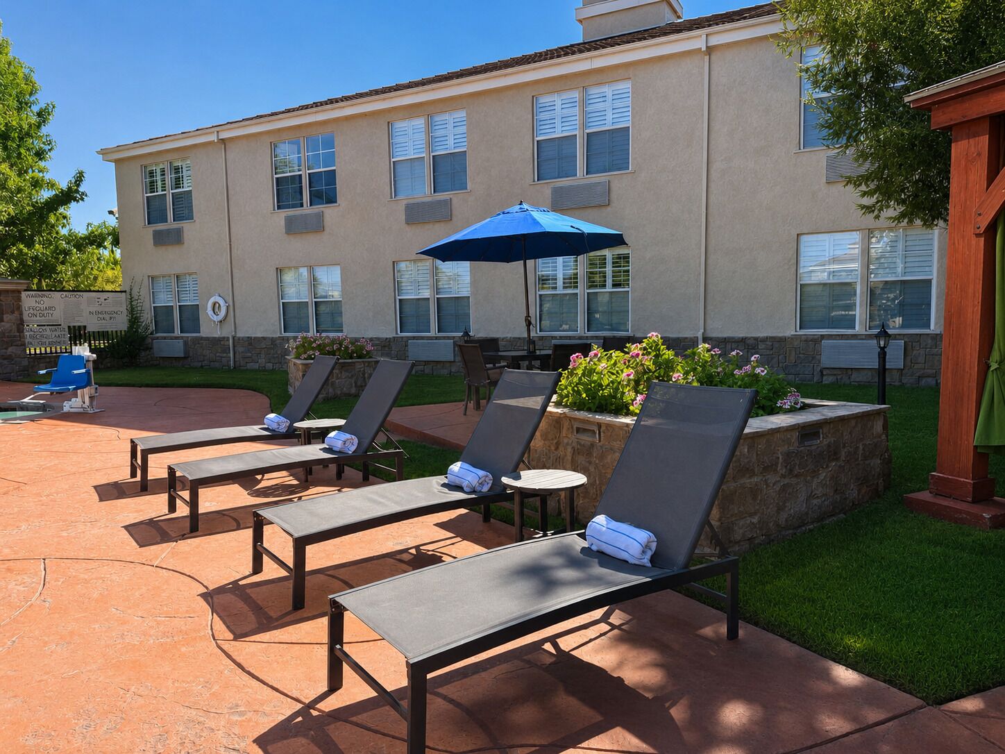 A patio area with lounge chairs and a table, a blue umbrella, and a building with multiple windows in the background.