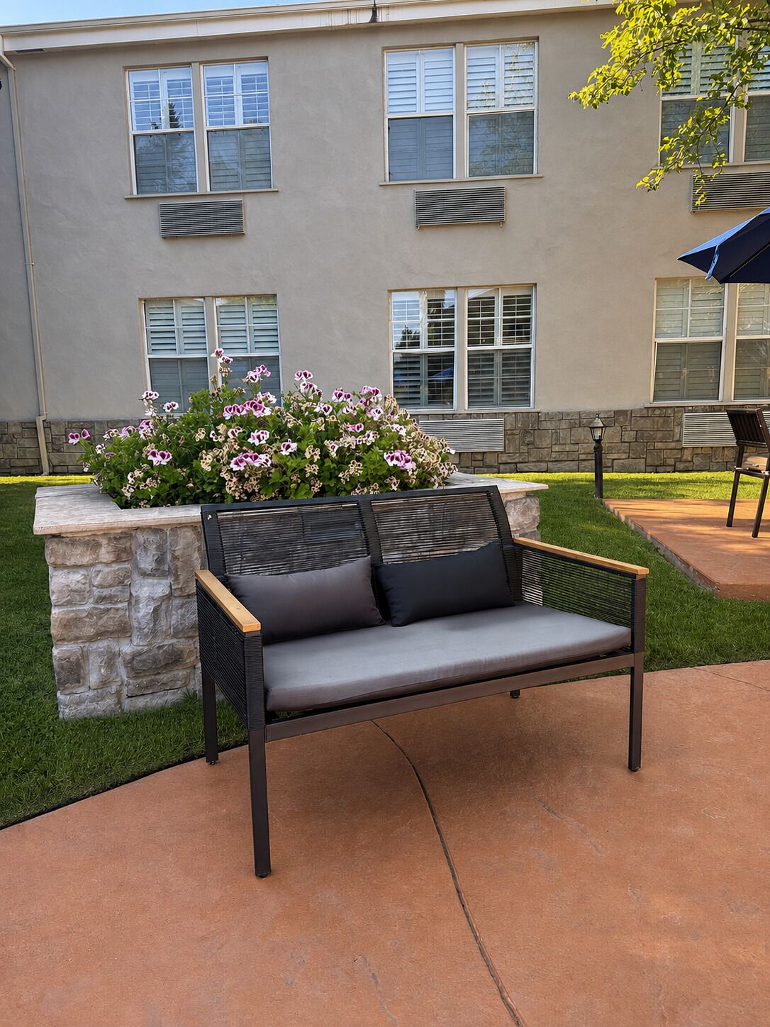 A patio area with a stone bench, a flower bed with pink flowers, and a building with multiple windows in the background.