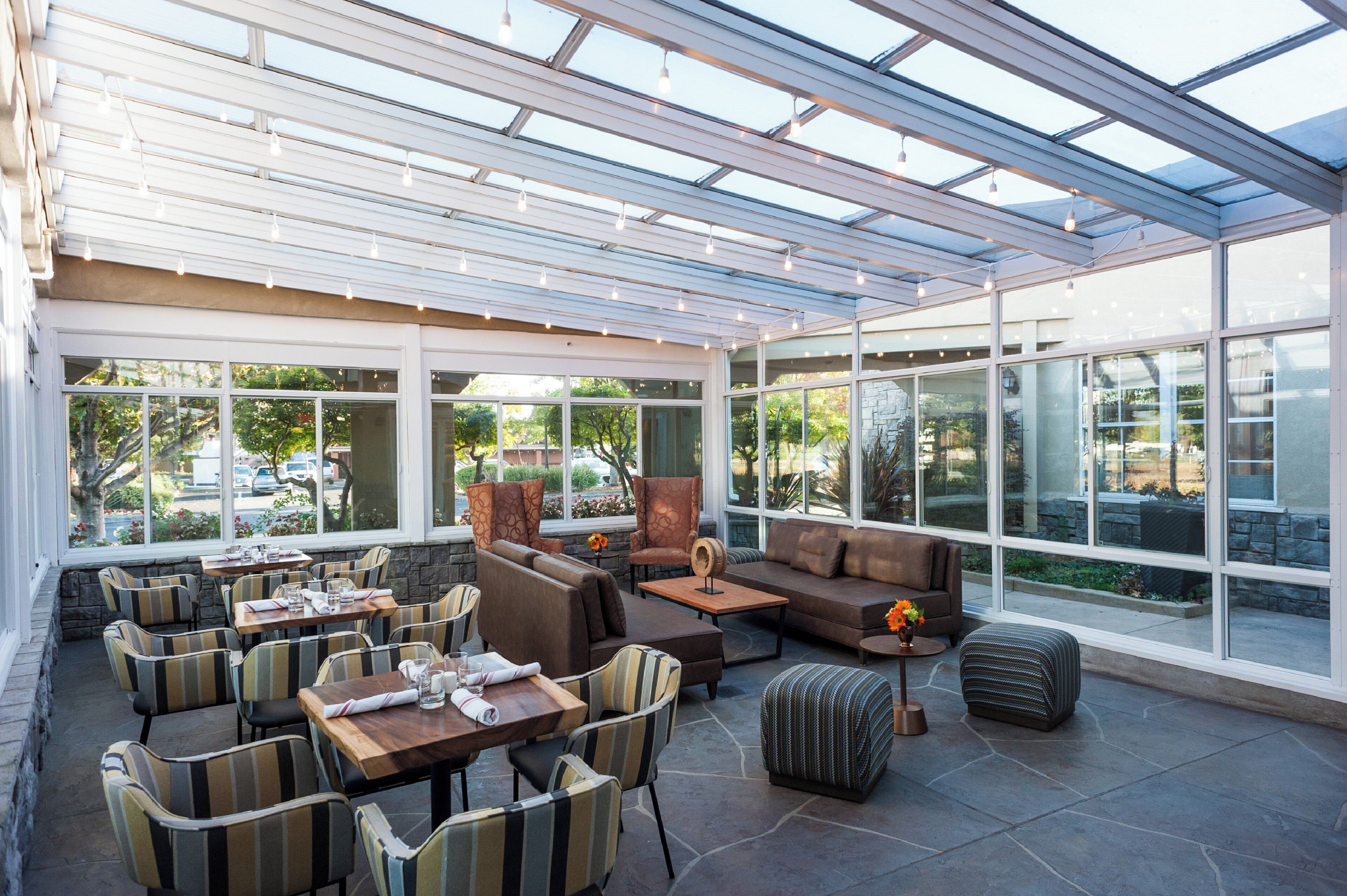 A sunlit indoor seating area with tables and chairs, large windows, and a glass ceiling.