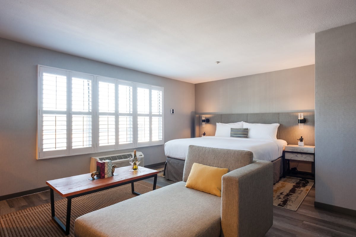 A hotel room with a large bed, a sofa, a small table with a bottle, and a window with blinds.