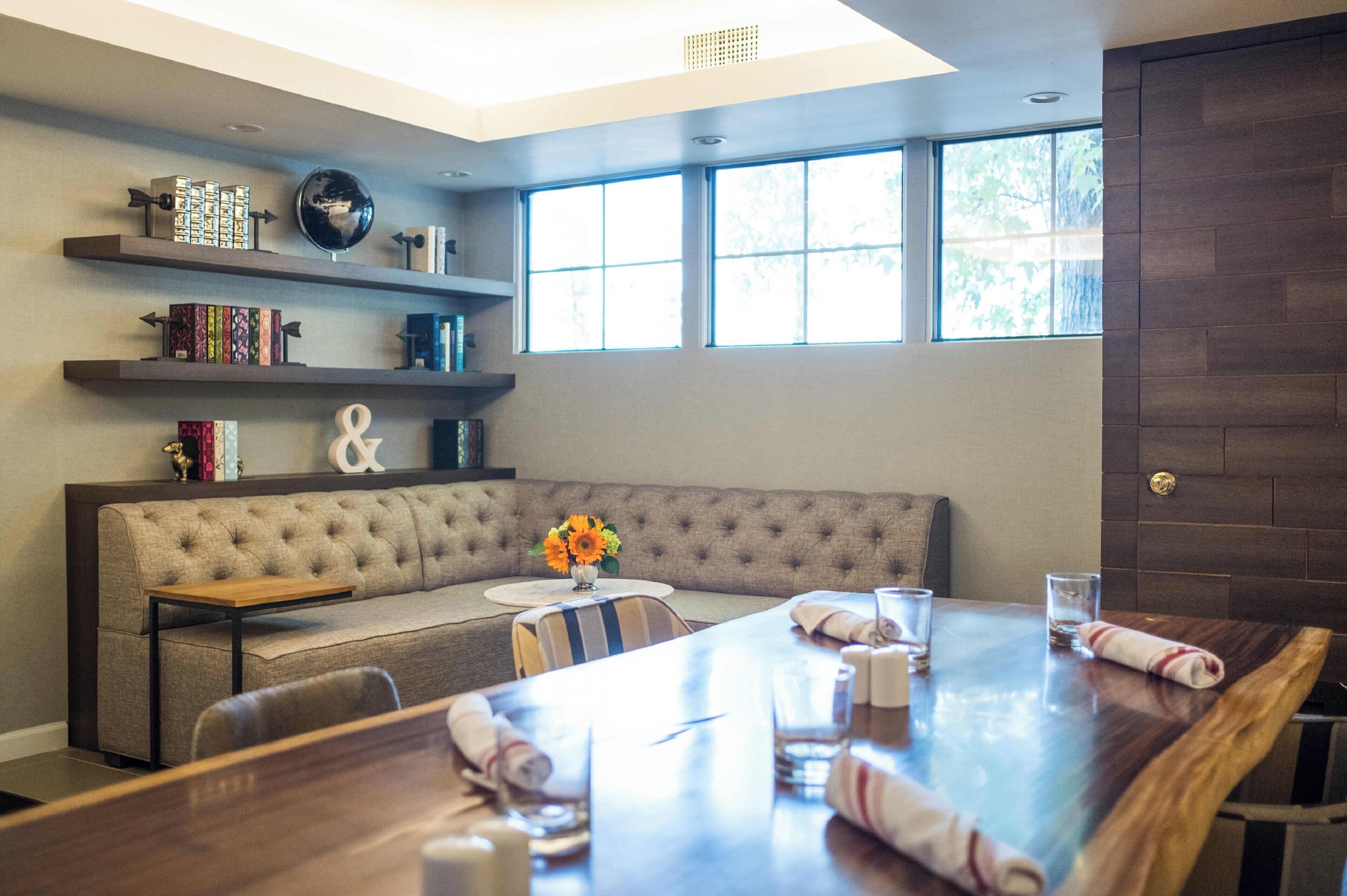 A modern living room with a sectional sofa, a dining table with chairs, a bookshelf, and a large window with natural light.